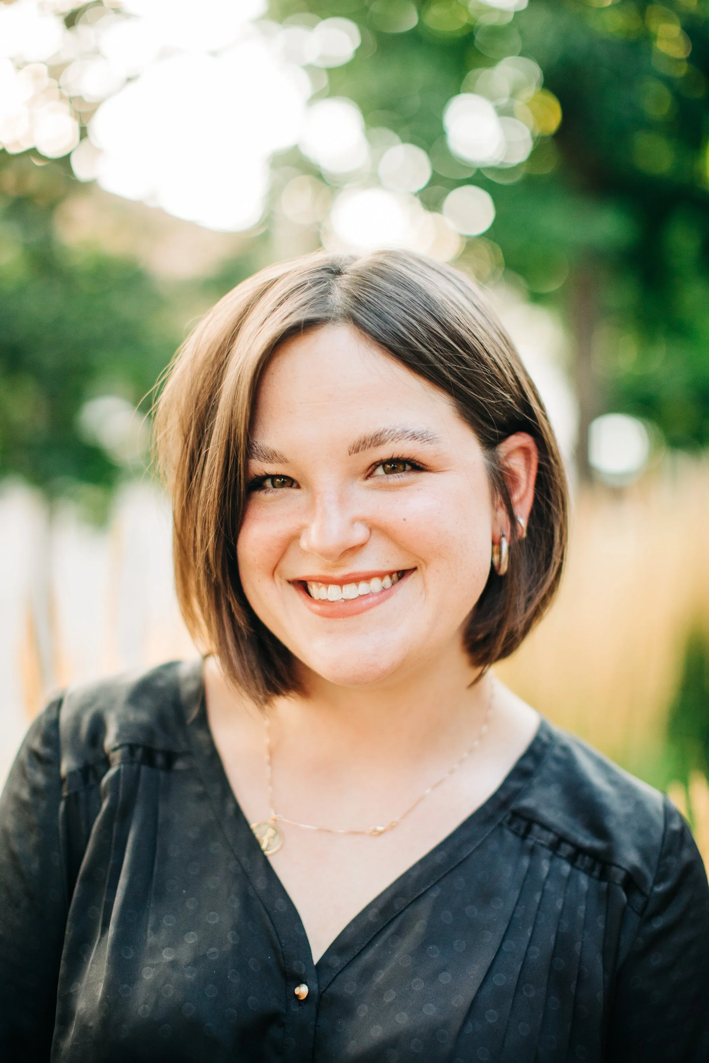 A smiling young woman with short brown hair wearing a black blouse and gold jewelry outdoors with a bokeh background of trees and sunlight.