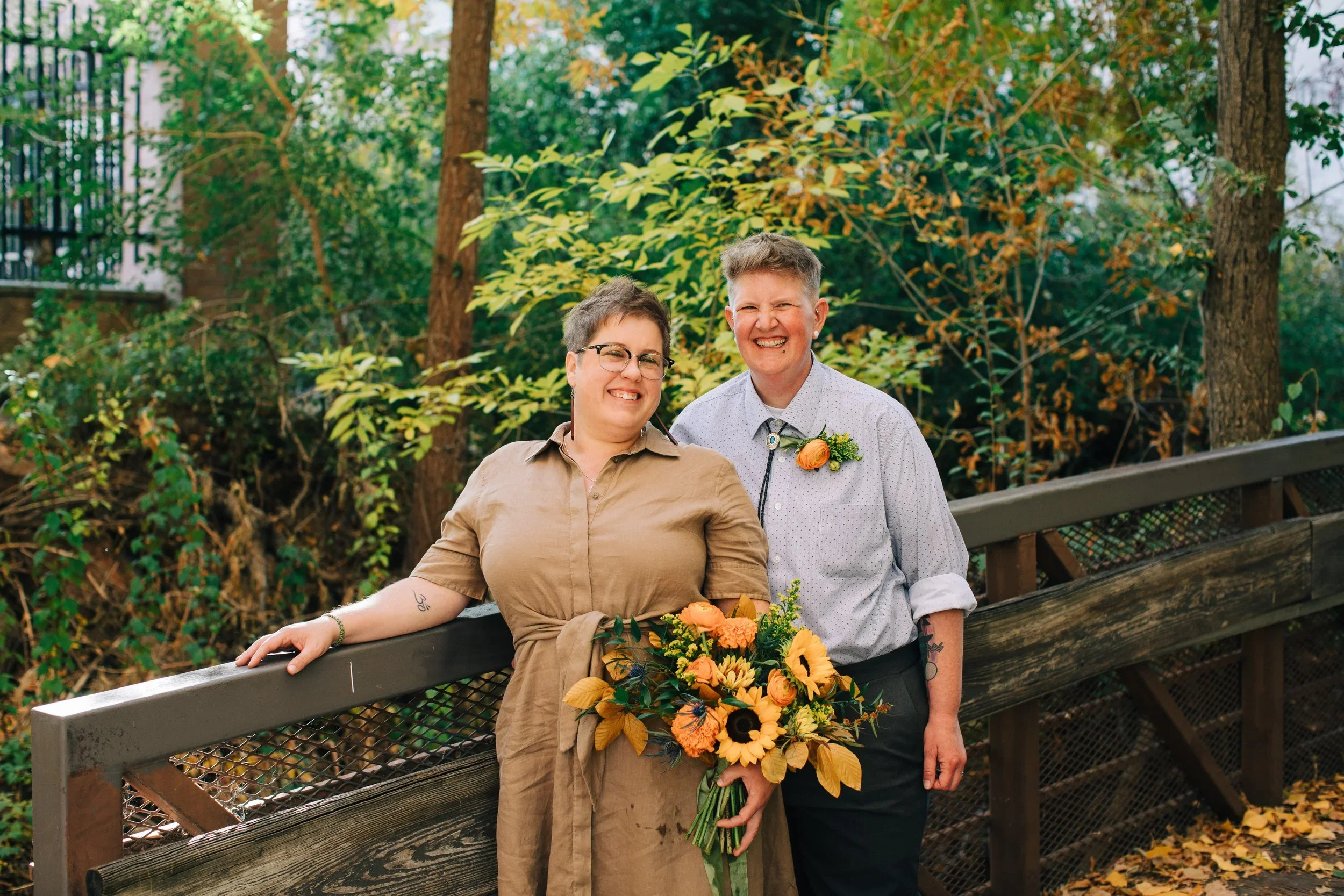 A queer and nonbinary couple is smiling and standing together outdoors, one holding a bouquet of yellow and orange flowers, surrounded by trees with green and yellow leaves, during autumn. One is masculine leaning, one is feminine leaning.
