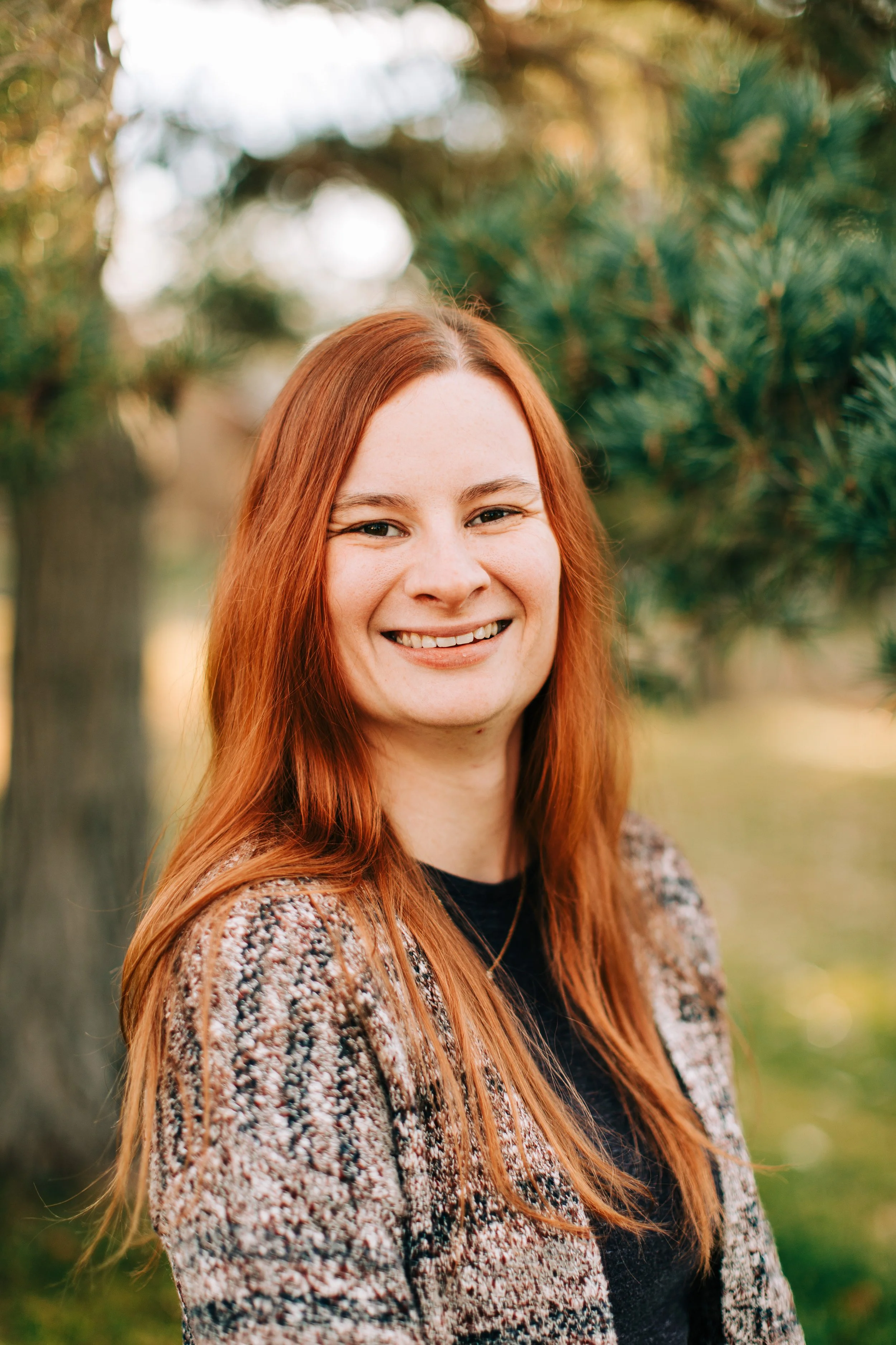 A smiling woman with long red hair standing outdoors near trees in a park in Montrose, Colorado.