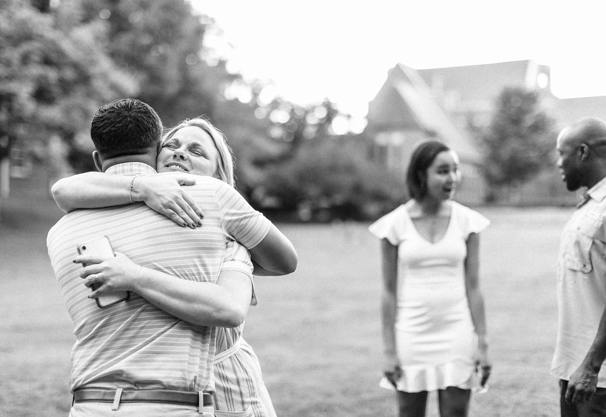A woman is hugging a man while holding a phone in her hand, and she appears happy. Two other people, a woman and a man, are standing apart in the background outdoors, on a grassy area with trees and houses in the distance.