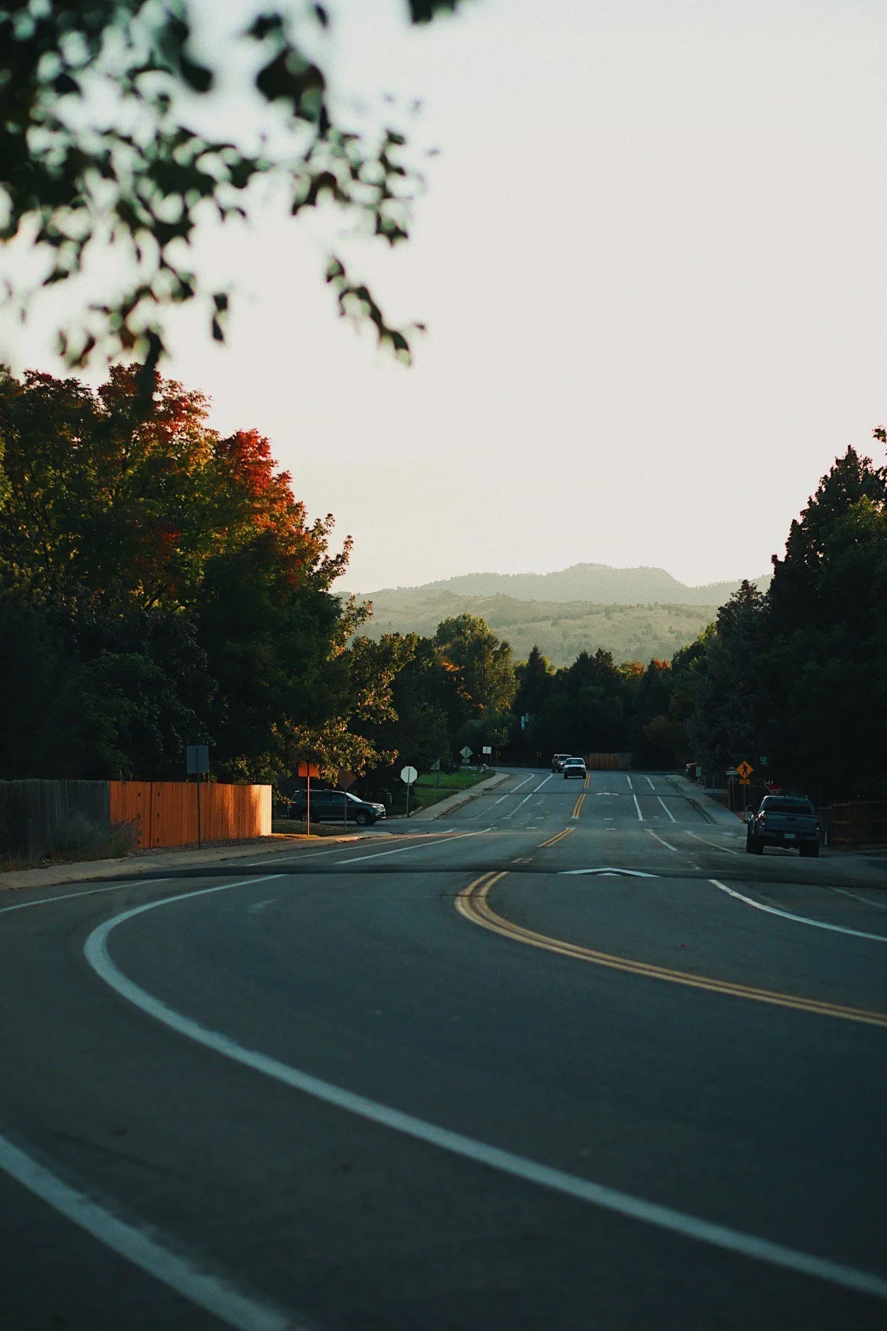 A winding two-lane road in a suburban neighborhood with parked cars and trees on both sides, leading towards distant mountains, during a time with soft sunset light.
