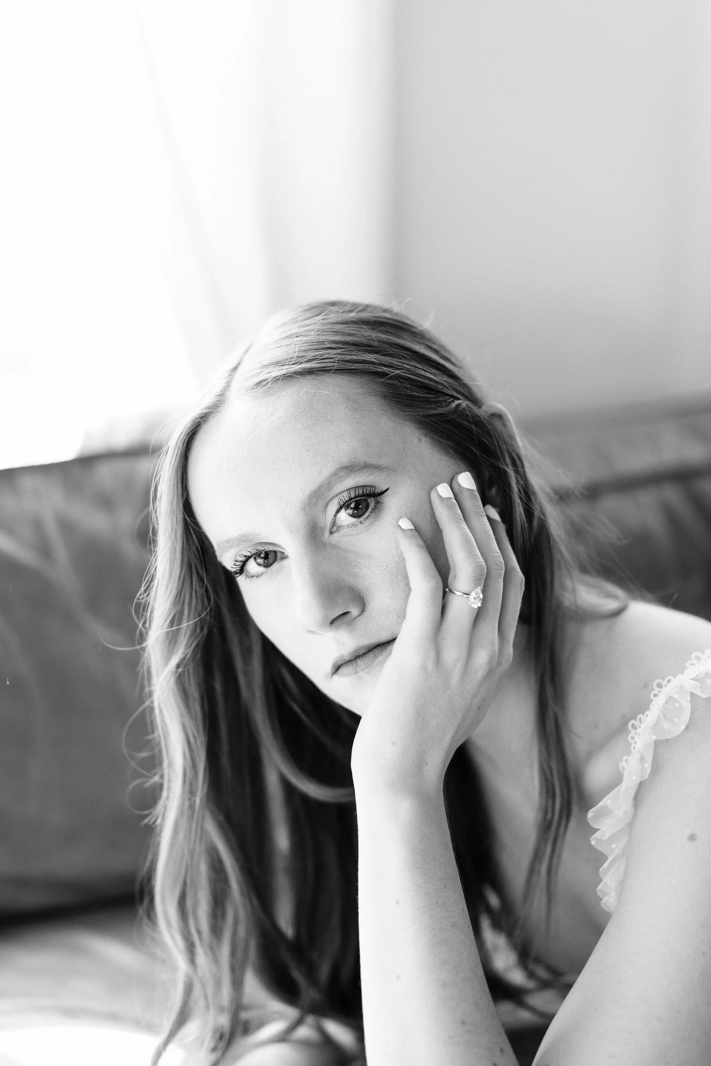 Black and white portrait of a young woman with long hair taken during a boudoir session, resting her face on her hand, wearing a ring, and looking at the camera.
