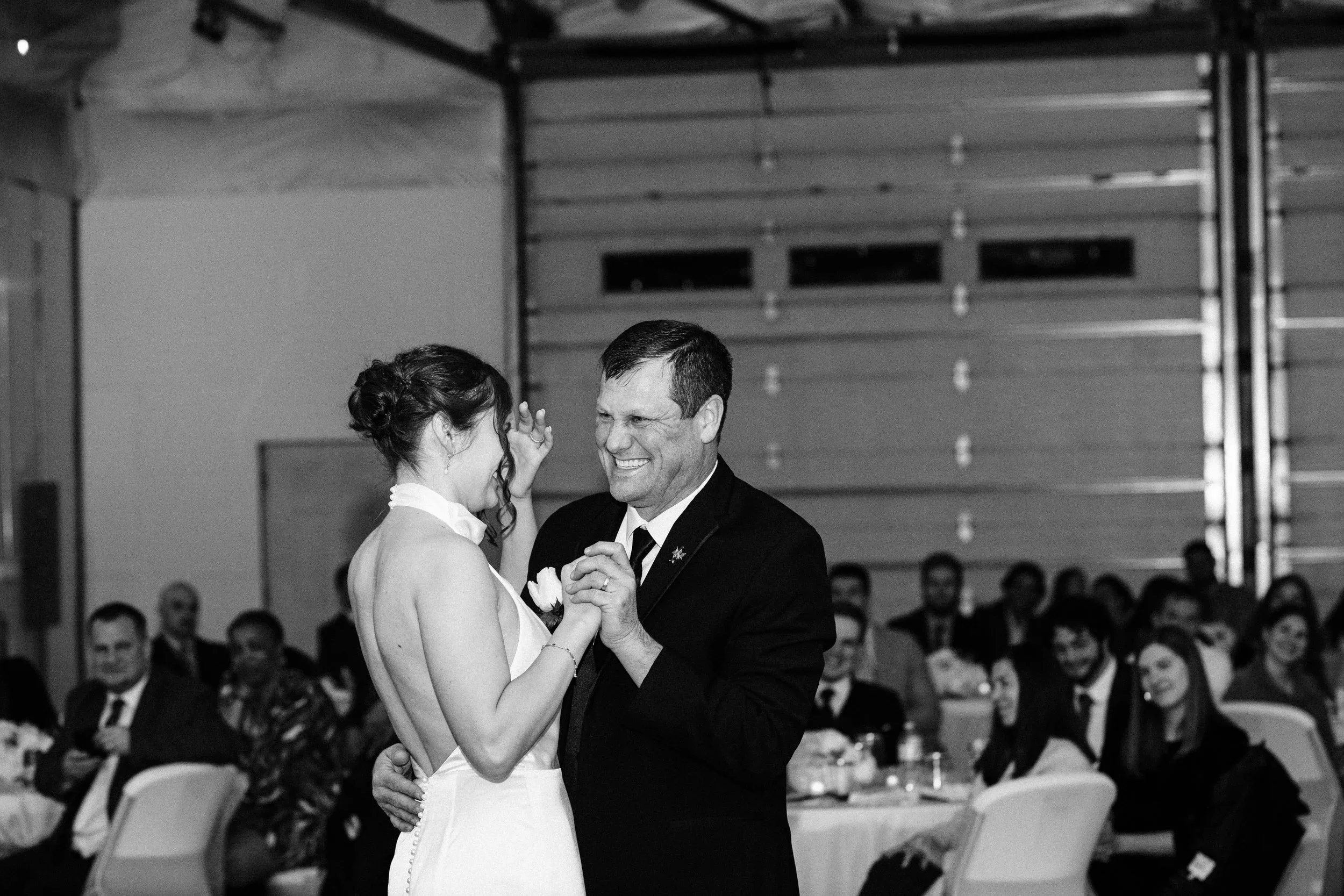 A black-and-white photo of a bride and an older man dancing at a wedding reception, with seated guests in the background.