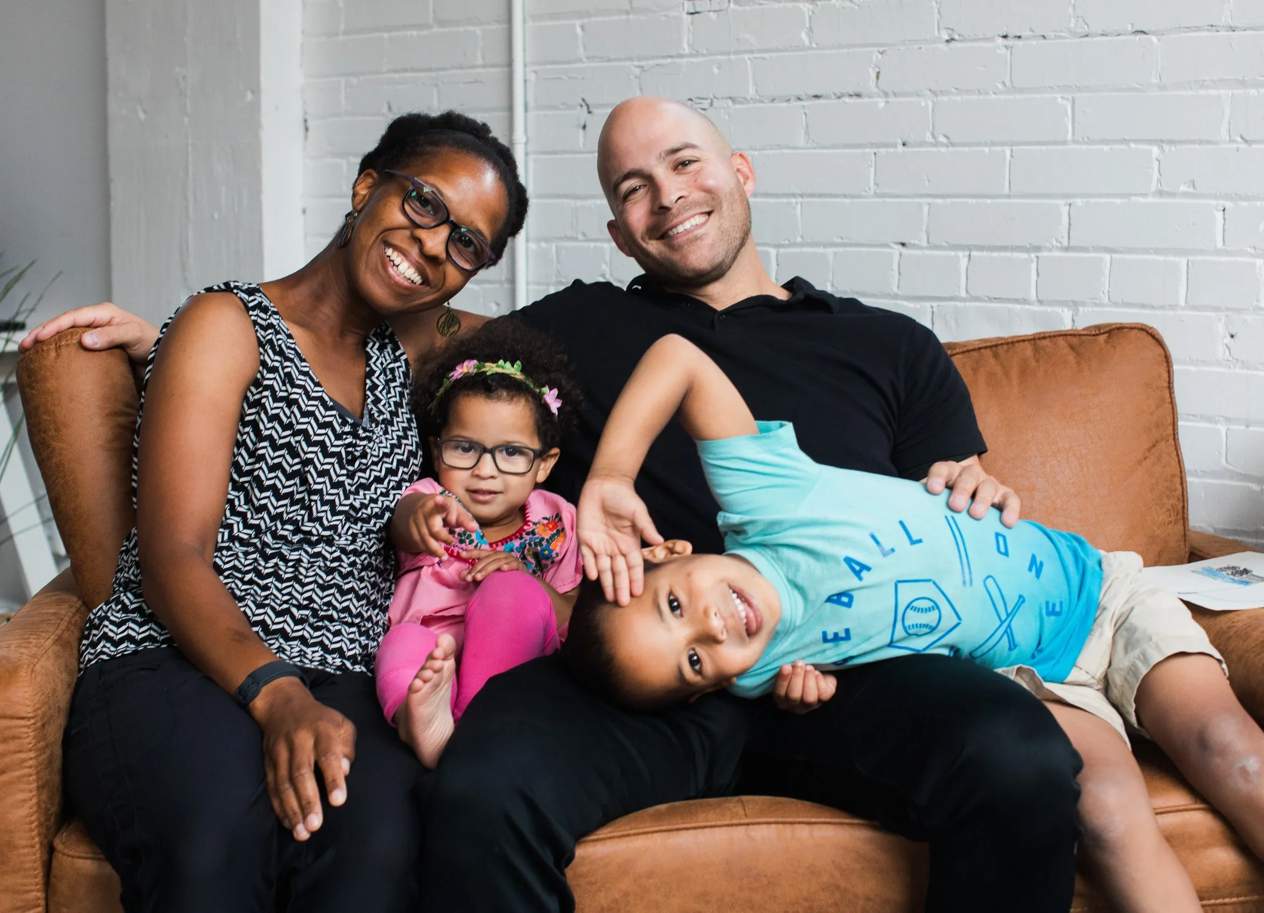 A diverse family of four sitting on a brown sofa in a room with white brick walls. The mother, wearing glasses and a patterned sleeveless top, is smiling. The little girl, with glasses and curly hair, is sitting on her father's lap, looking at the ca