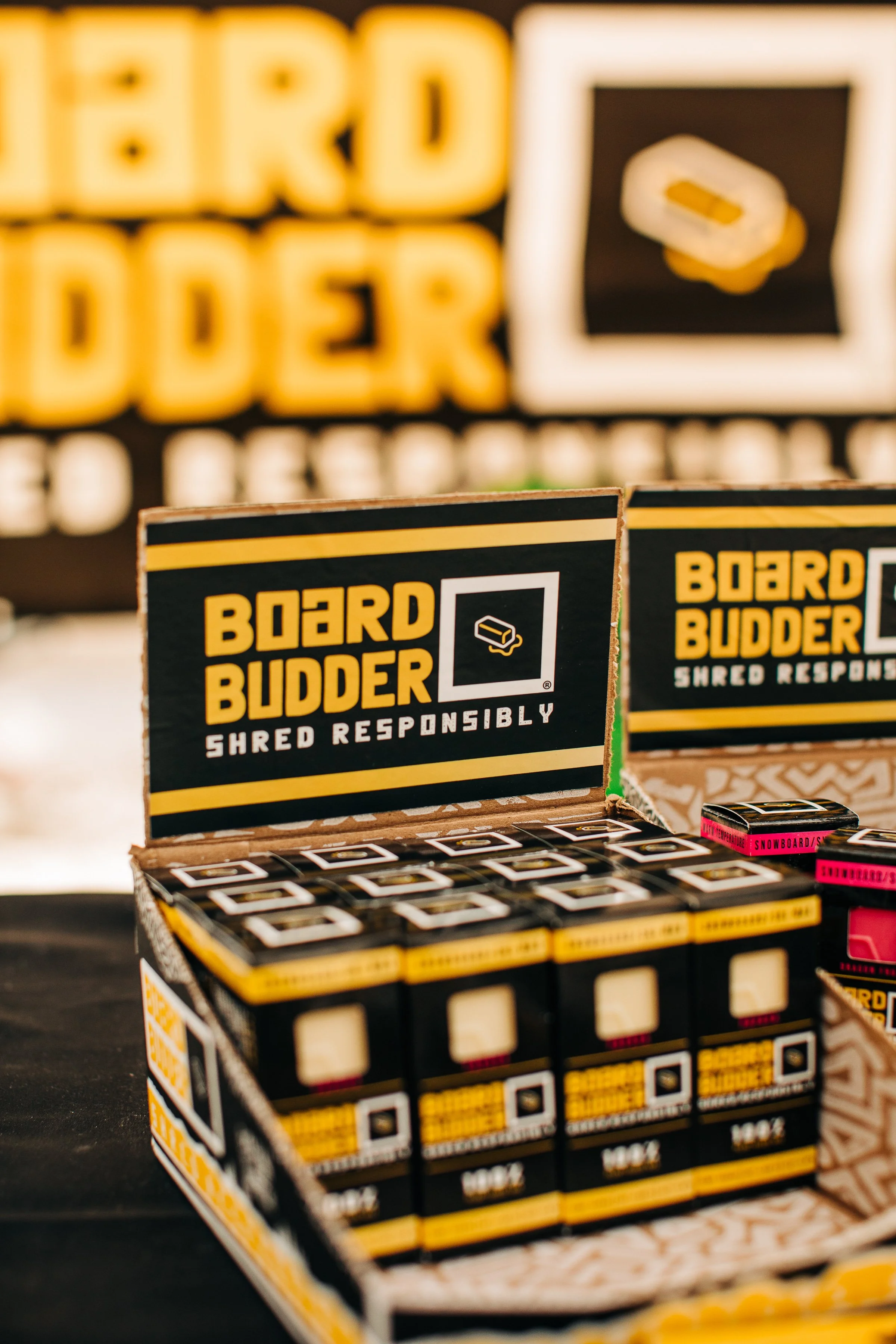 A display of Board Budder product boxes with black, yellow, and white packaging, featuring the logo and the slogan 'Shred Responsibly' taken at a Brighton UT nonprofit festival