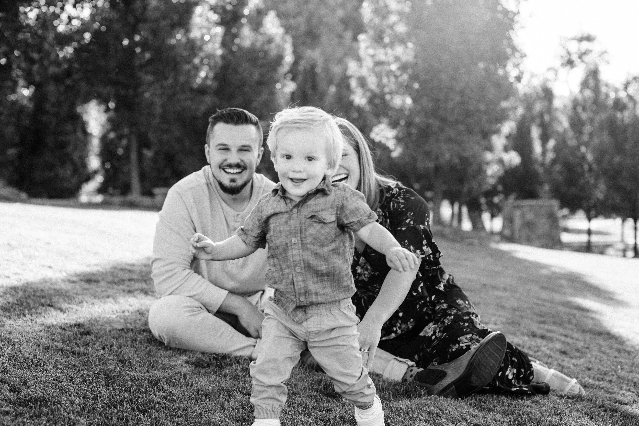 A family of three sitting on grass in a park, smiling at the camera, with trees in the background. Spring Valley Park in Fort Collins, CO. The focus is on a toddler standing in front, with a man and woman behind him, all smiling.