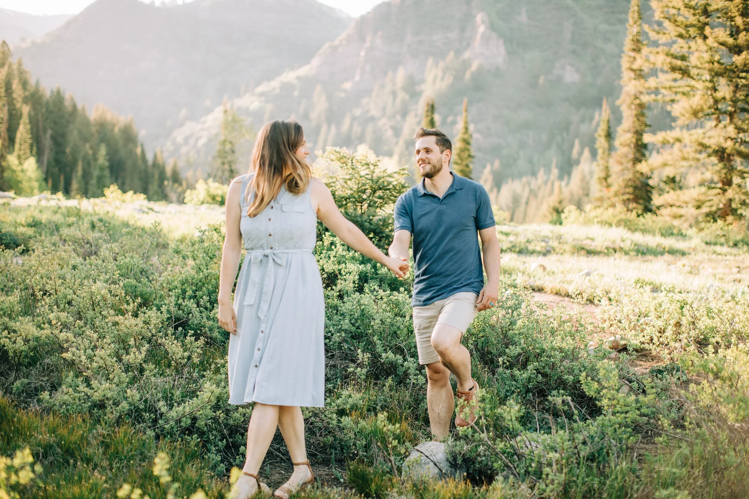 A couple walking hand in hand through a forested mountain area during sunset, smiling at each other. The setting is Jordan Pines natural area in Utah.