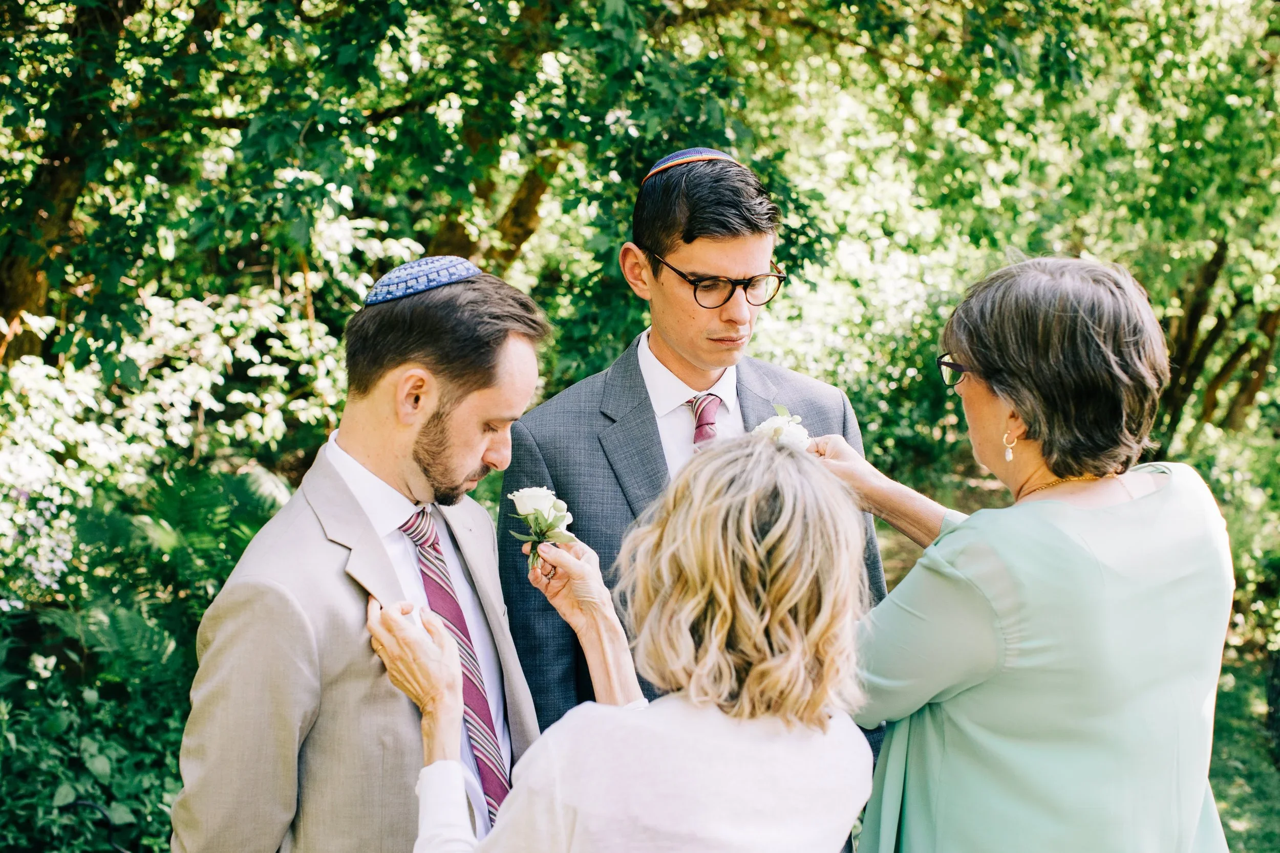 A group of people participating in a Jewish wedding ceremony outdoors, with a rabbi pinning a white flower boutonniere on a groom's suit. The groom and another man are wearing yarmulkes, and the scene is surrounded by green foliage.