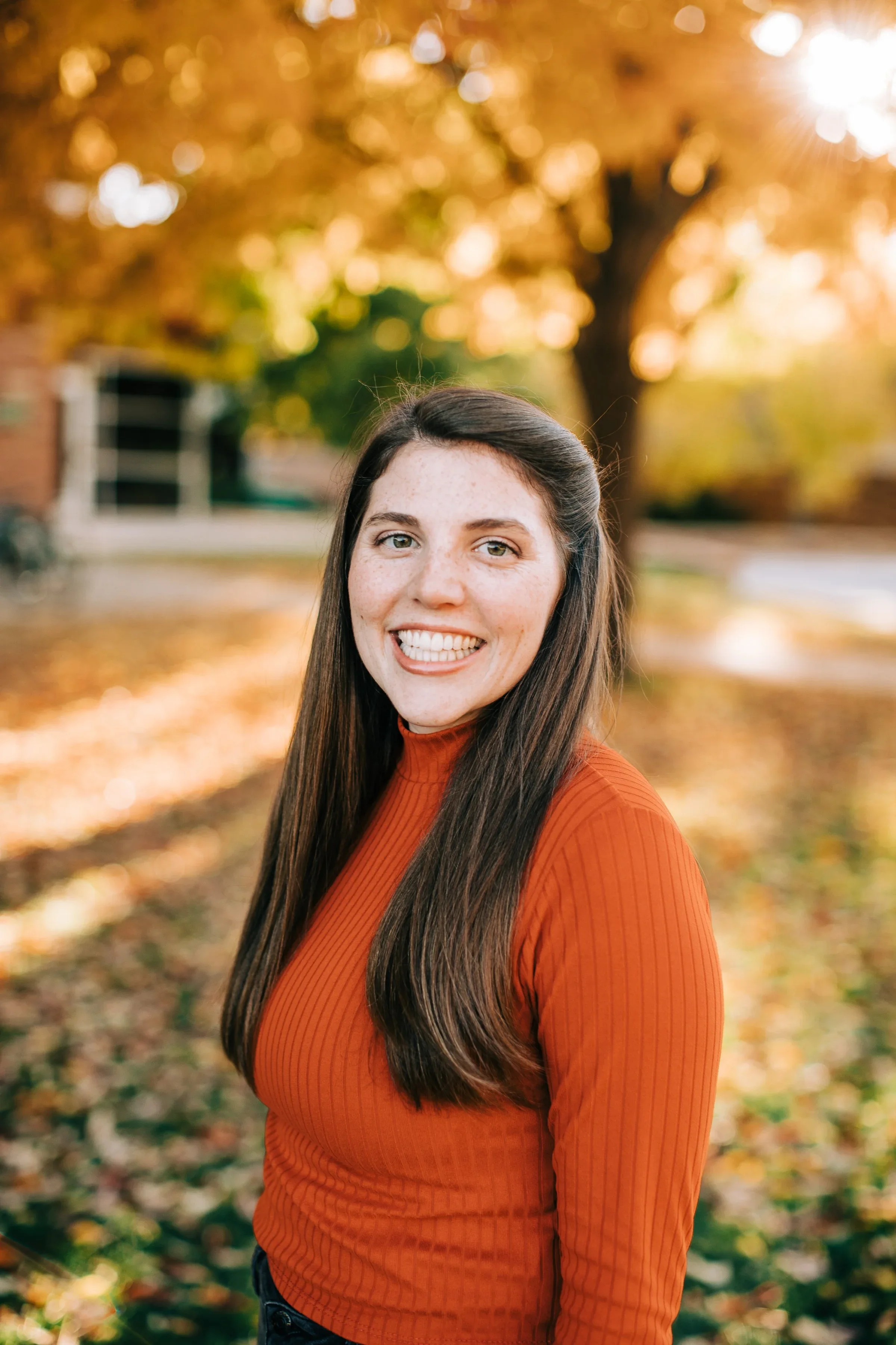 A smiling woman with long brown hair and freckles standing outdoors during fall, with trees with orange and yellow leaves in the background.