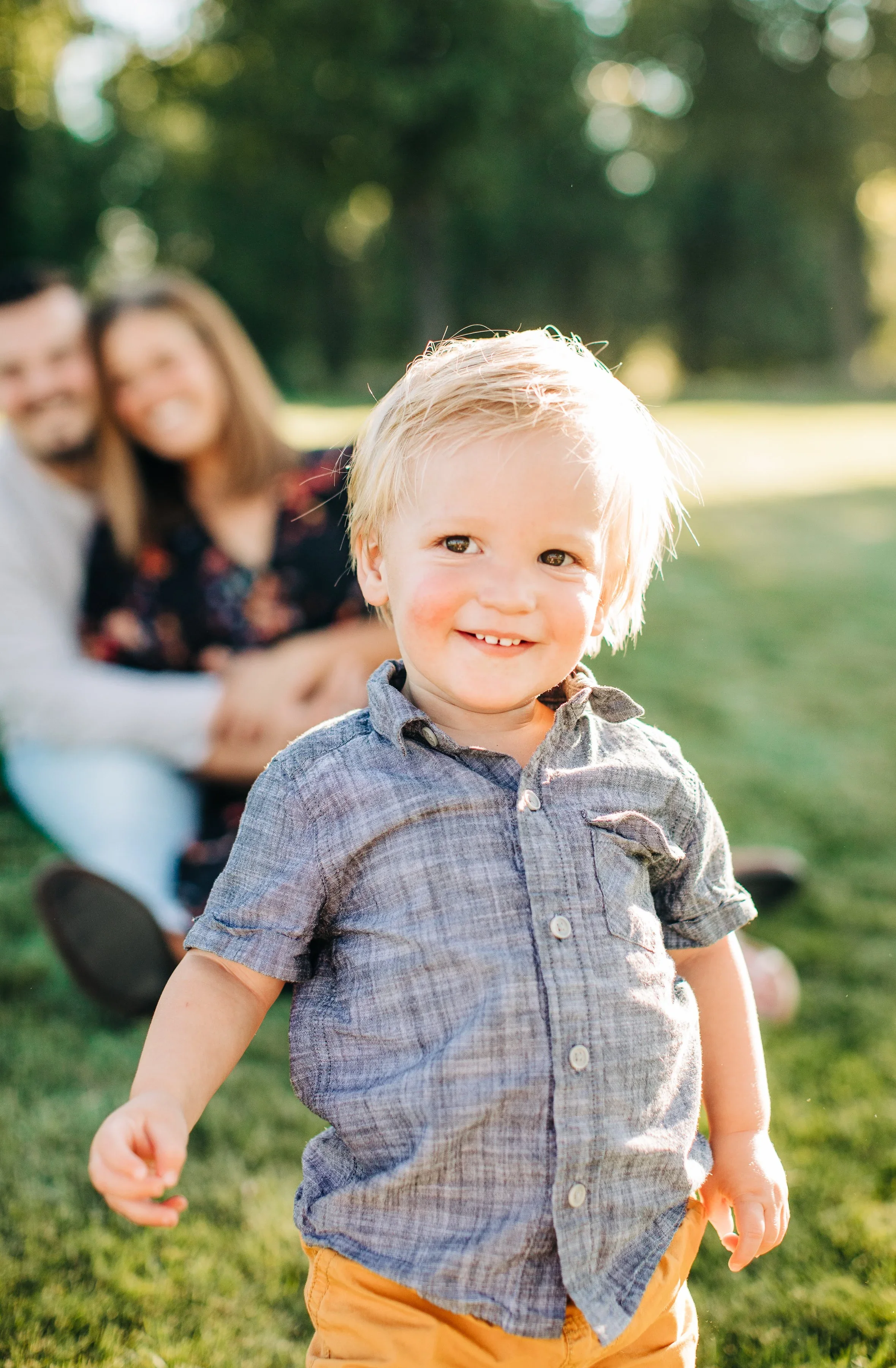 A young boy with blonde hair and a wide smile standing outdoors on grass; a blurred couple, his parents (Fort Collins couple) are sitting on the grass behind him.