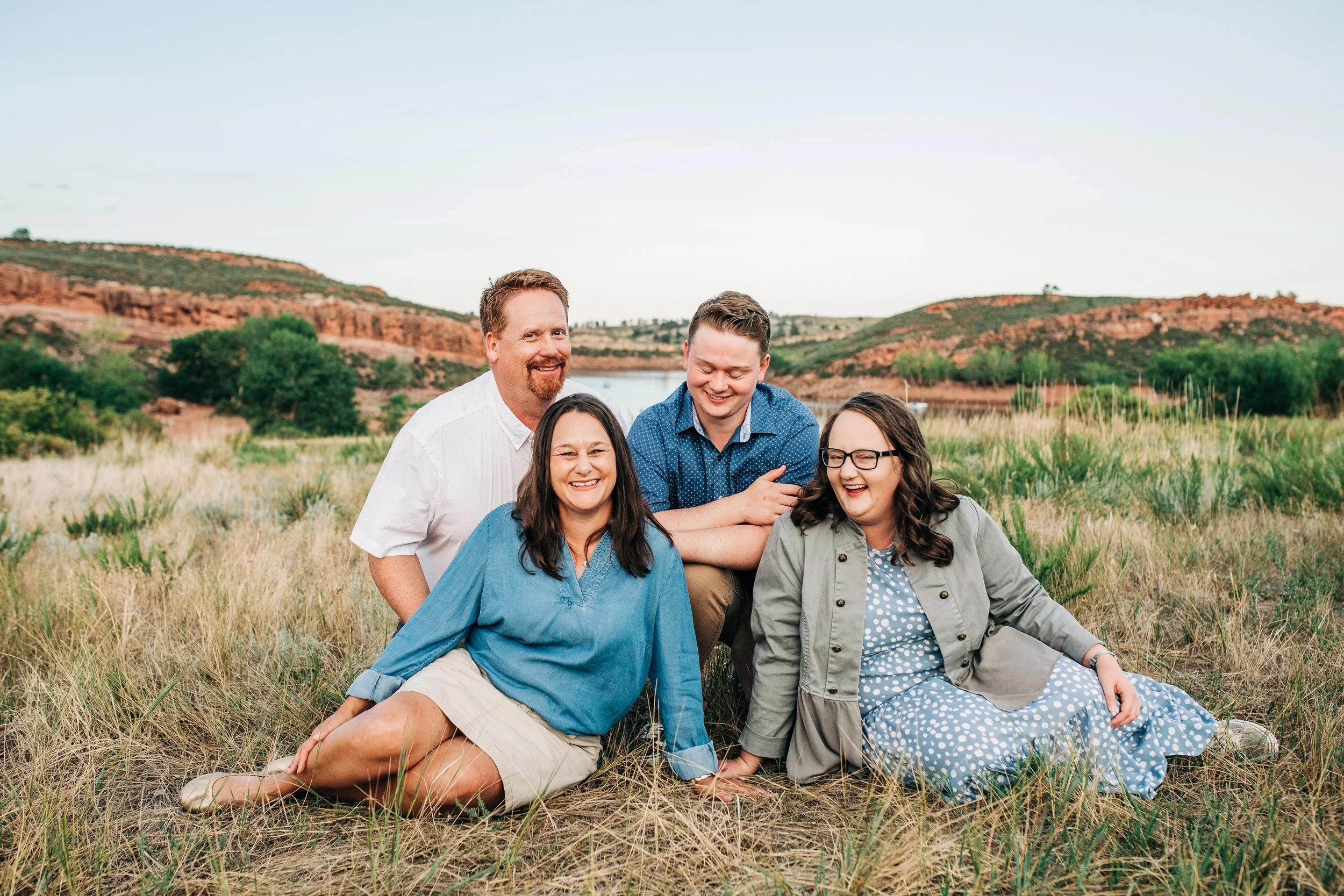 A group of five people sitting and standing in a grassy field with a river and red hills in the background. They are smiling and laughing.