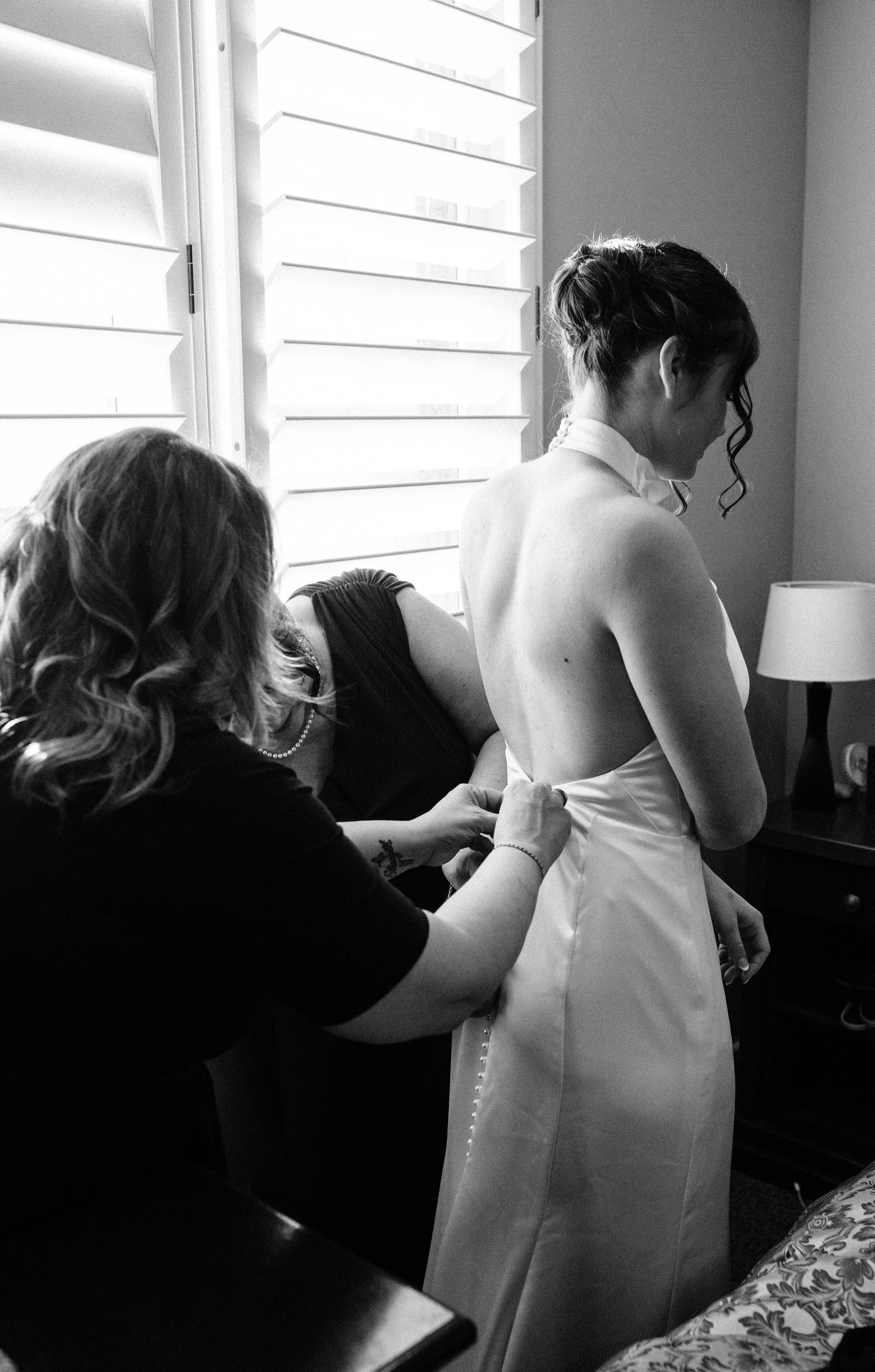 A woman in a white dress is being helped into her dress by another woman in a black dress. The scene takes place near a window with shutters.