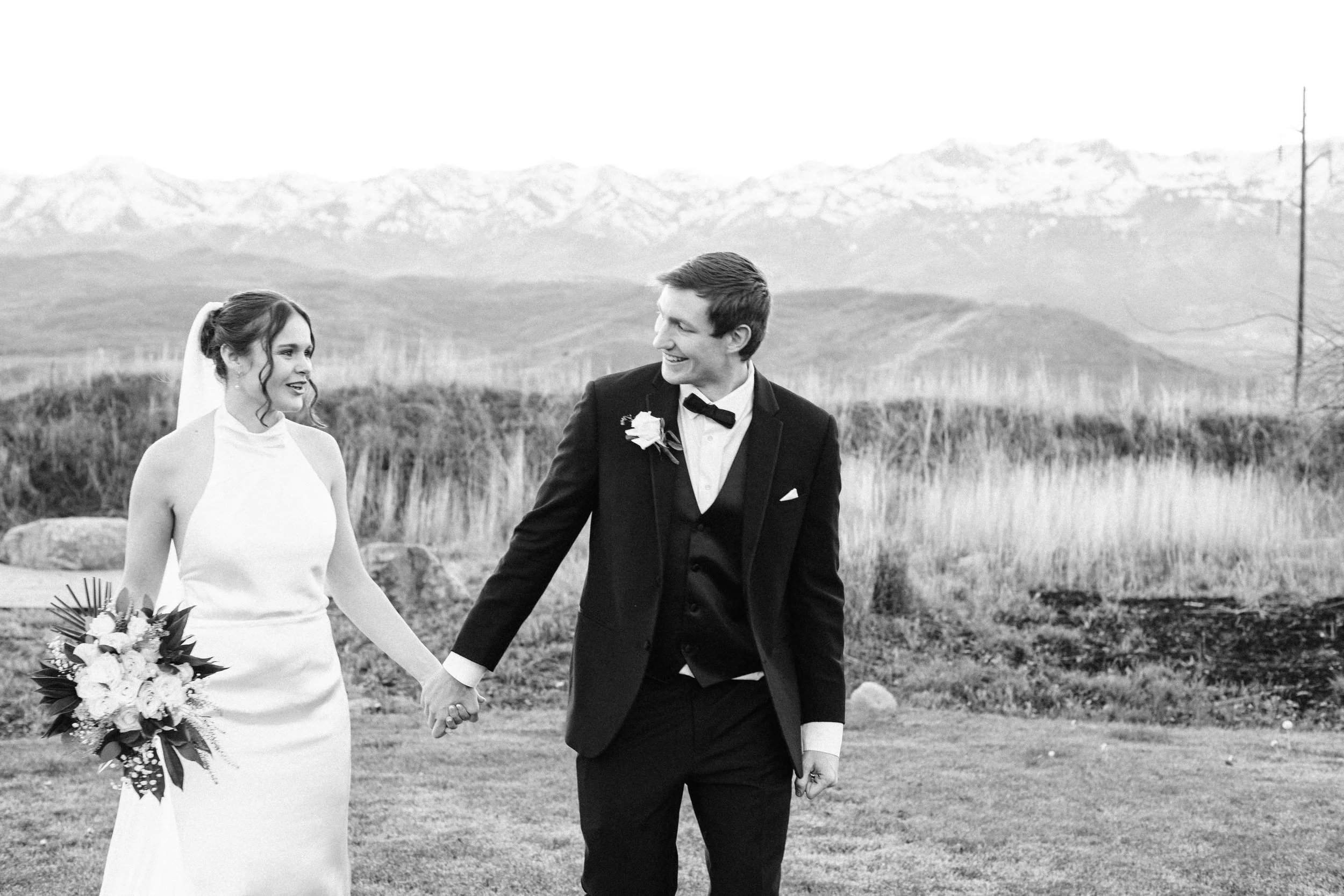 A bride and groom holding hands outdoors, with the Wasatch mountains in the background, engaged in a wedding ceremony or photoshoot.