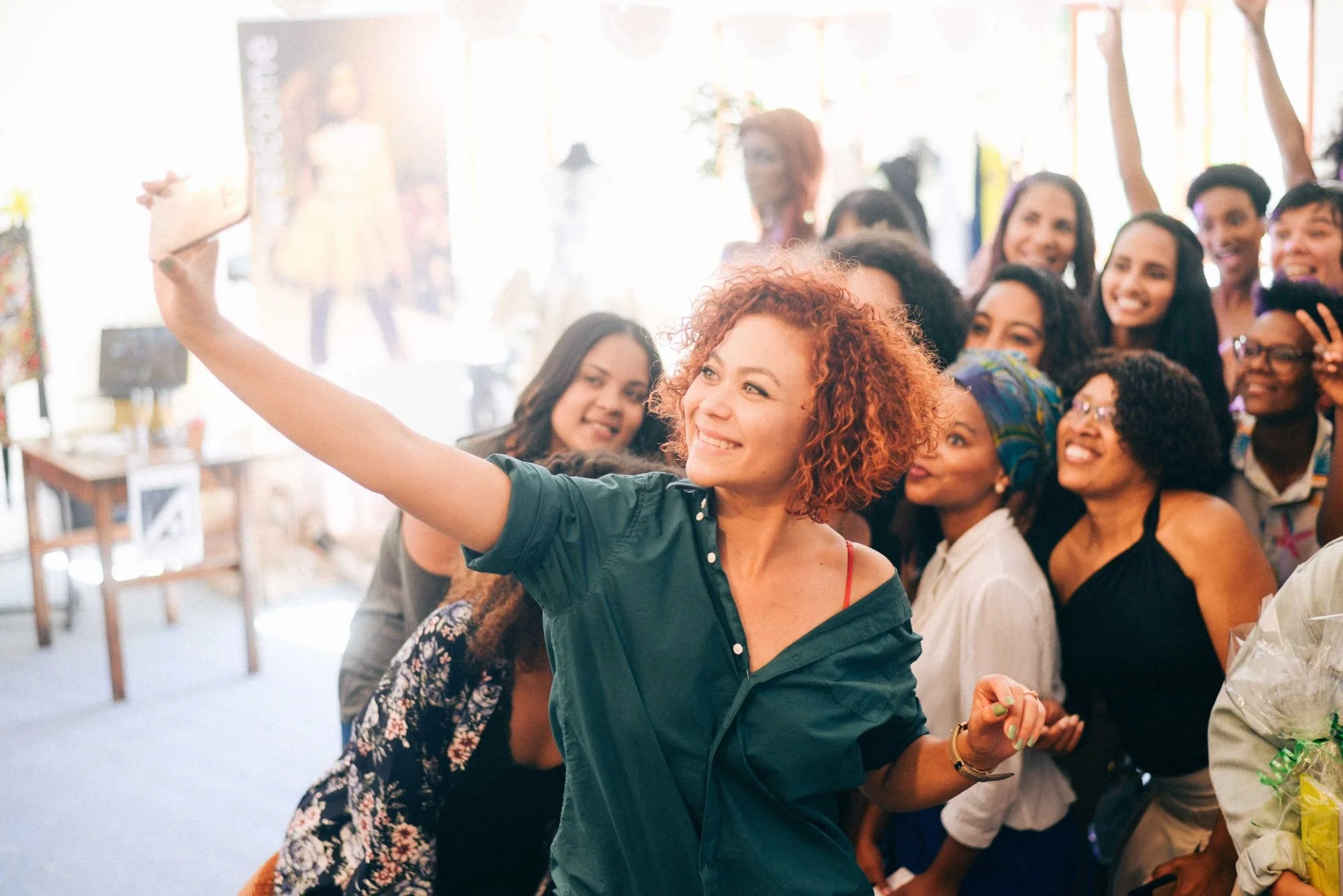 A group of diverse women taking selfies and celebrating together at an indoor event with bright natural lighting.