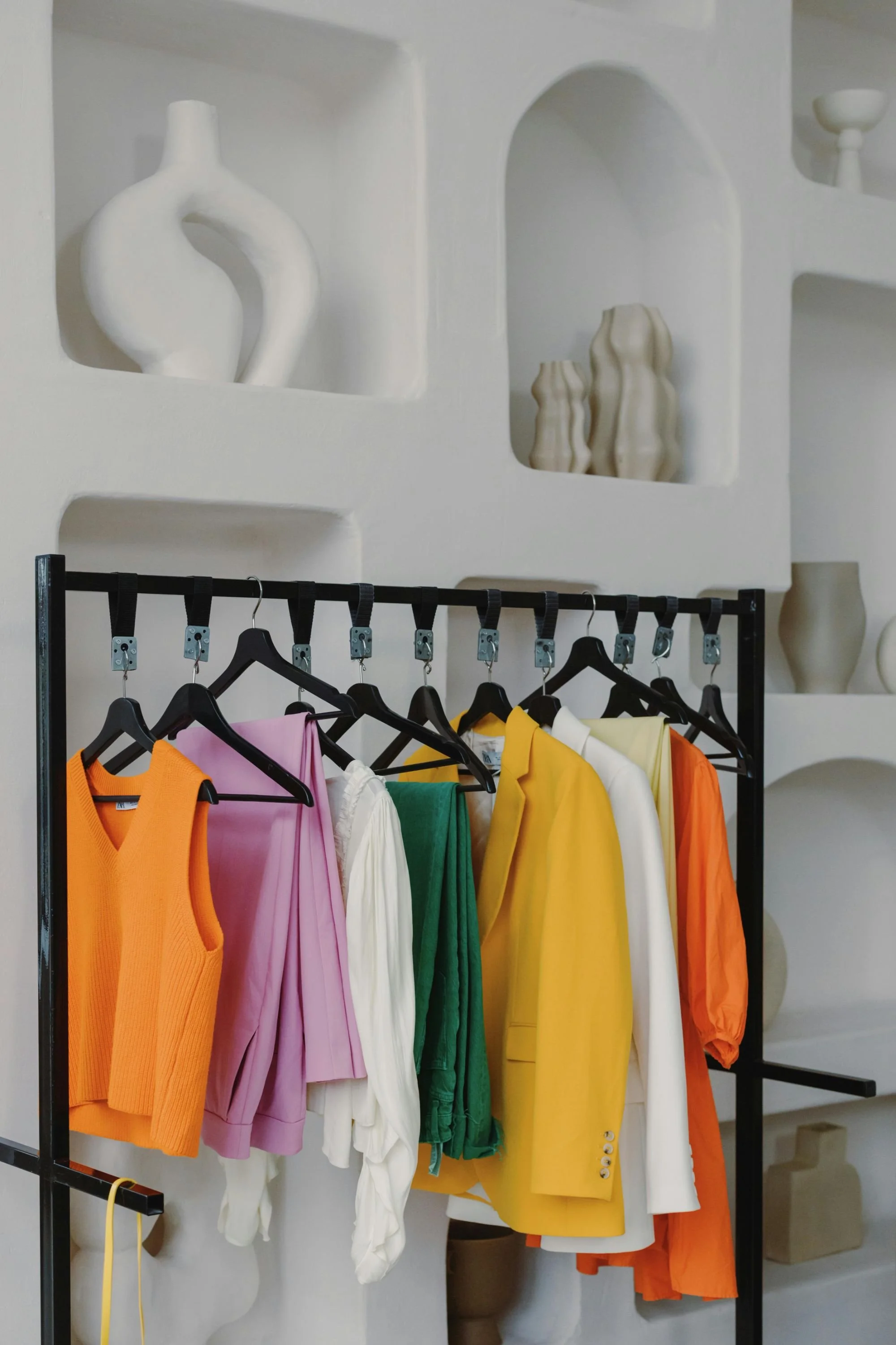 Colorful sleeveless tops and dresses hanging on black clothing rack with black hangers in a clothing store. Decorative white vases on white wall shelves in the background.