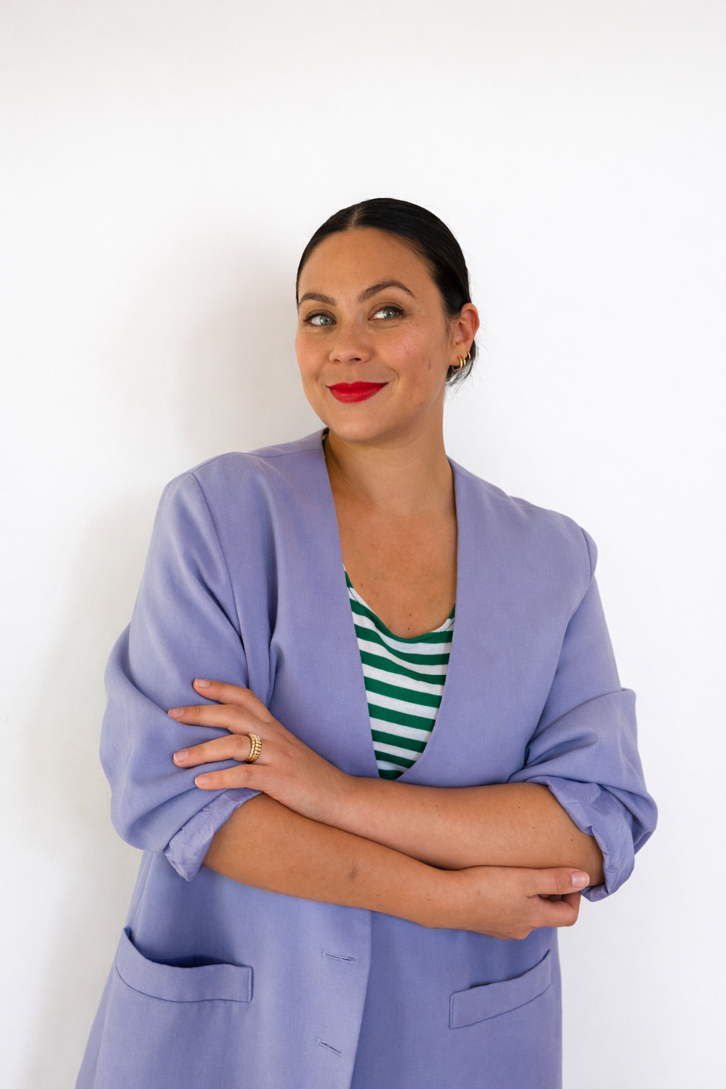 A woman with dark hair, red lipstick, and earrings standing against a white wall, wearing a purple blazer over a green and white striped top, with her arms crossed.