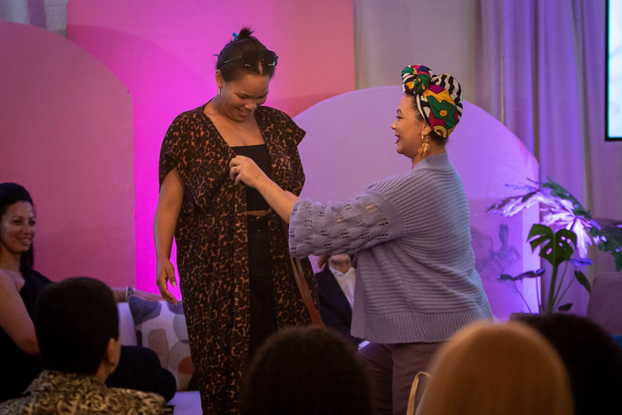 Two women are on stage at an event, one wearing a leopard print dress and the other in a gray sweater with a colorful headwrap. They are smiling and engaging, with the woman in gray pinning a flower to the other woman's dress. Several people are seated nearby, watching the interaction.