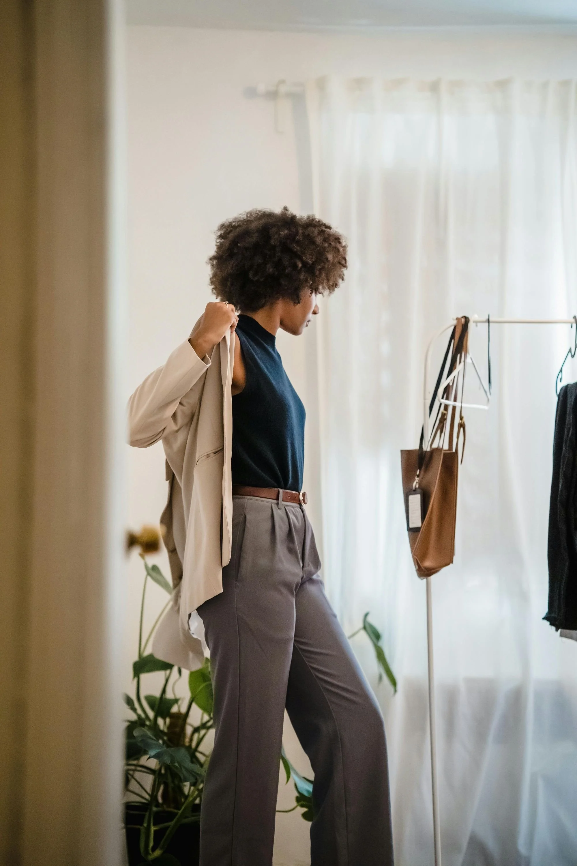 A woman with curly hair, wearing a navy top, beige blazer, and gray pants, standing indoors near a clothing rack with bags and clothing, and a green plant nearby, opening her beige blazer.