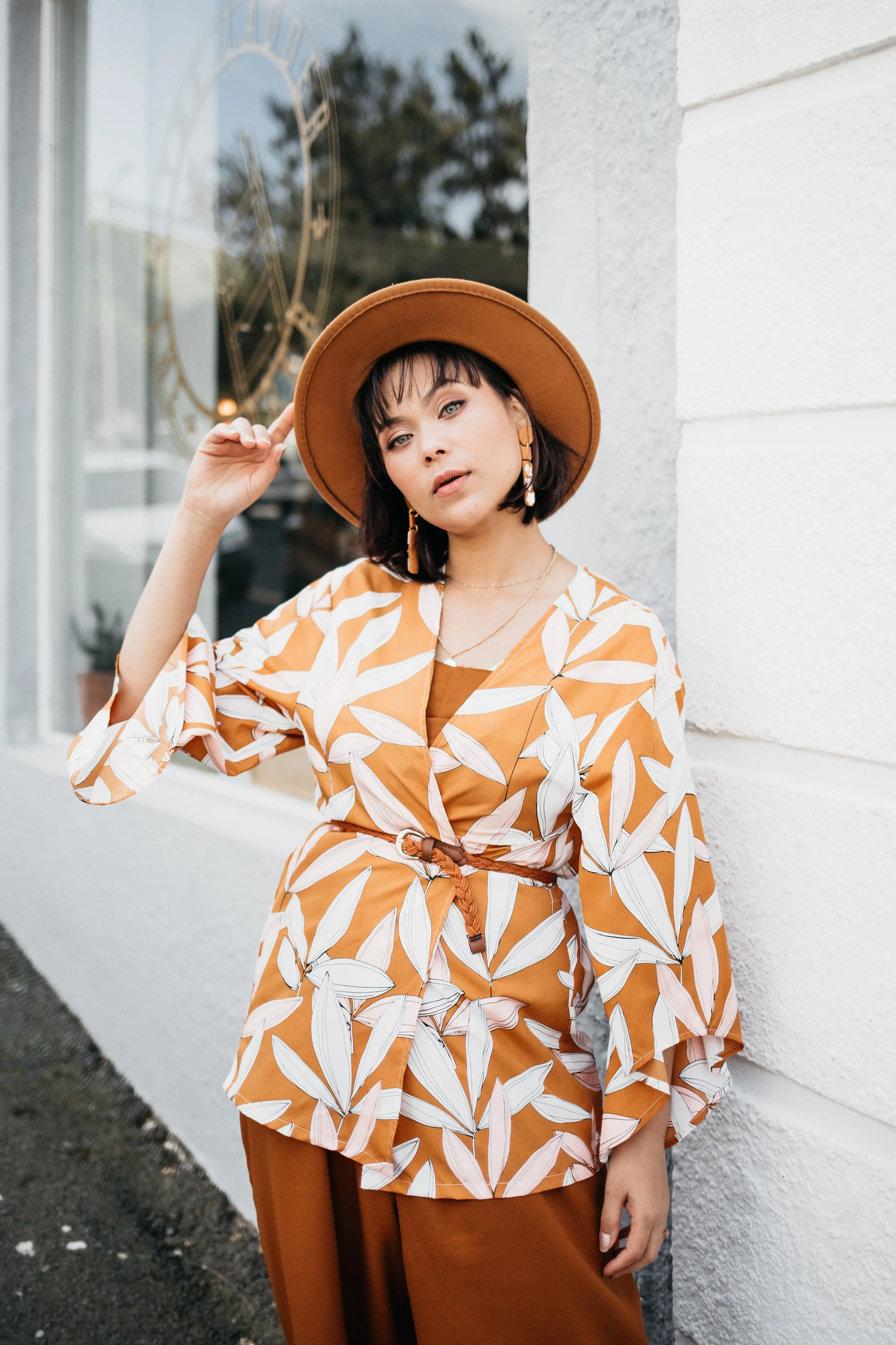 A woman wearing a wide-brimmed tan hat and a matching orange and white floral-patterned blouse poses outside near a white wall, with a clock visible through the window behind her.