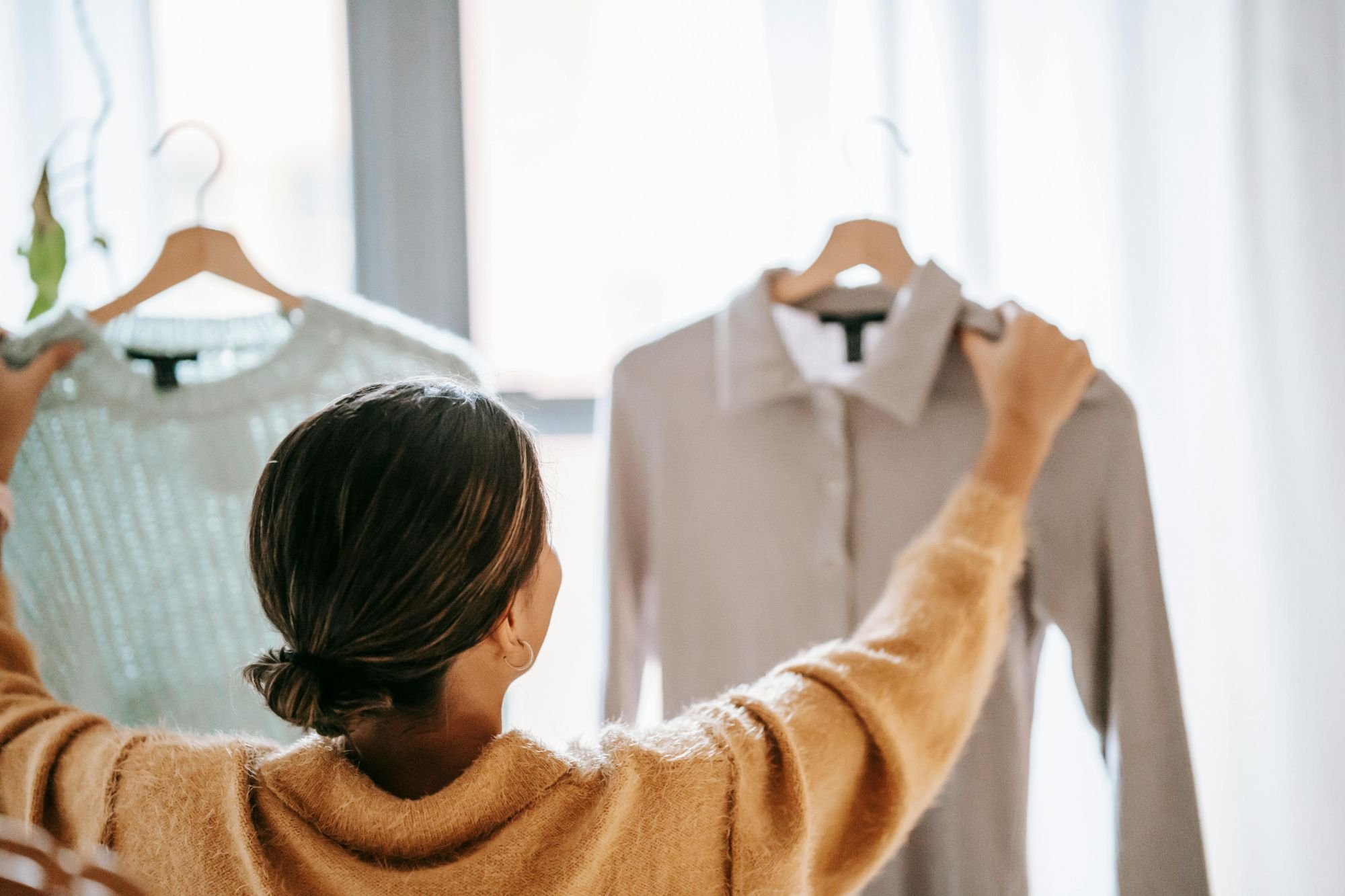 A woman with dark hair tied back holds up a white shirt on a hanger, looking at it in front of a window with sheer curtains, while another woman, partially visible, holds a similar shirt on a hanger.