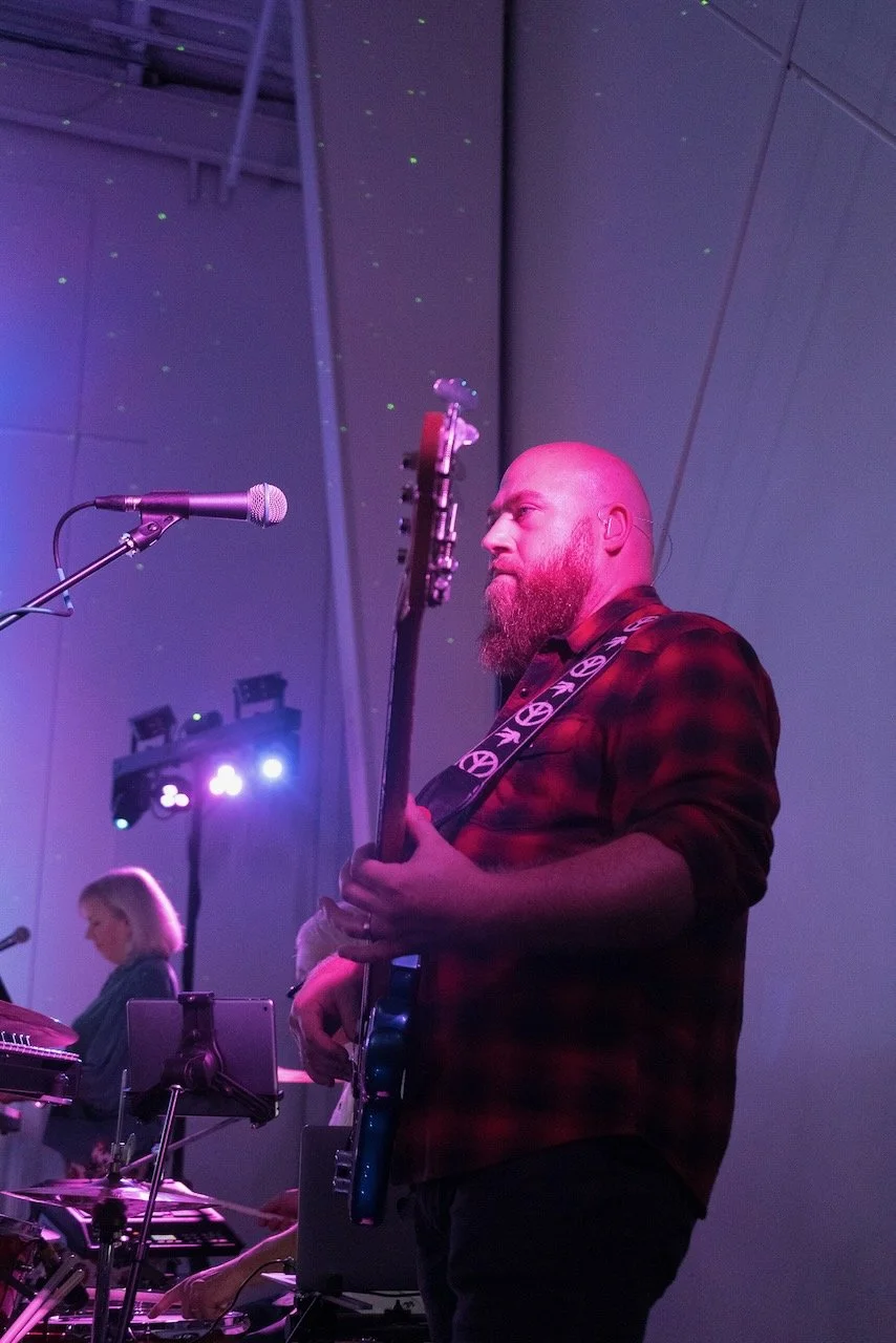 A man with a beard playing an electric guitar on stage under purple and blue lighting, with a microphone nearby and other musicians in the background.