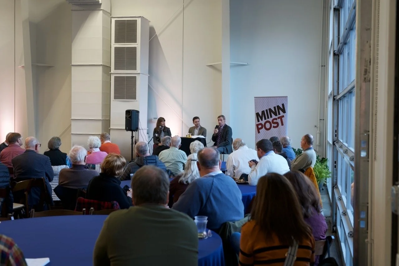 A panel discussion at a conference with three speakers seated at a table on a stage, in front of an audience in a large, well-lit room with tall windows. A banner with 
