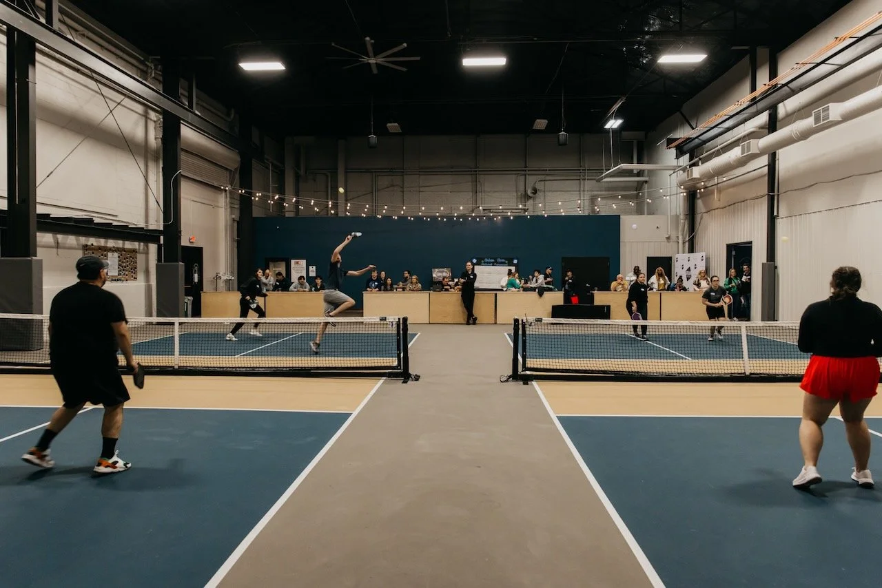 Indoor pickleball game with players on two courts, spectators watching, and a bar area in the background.