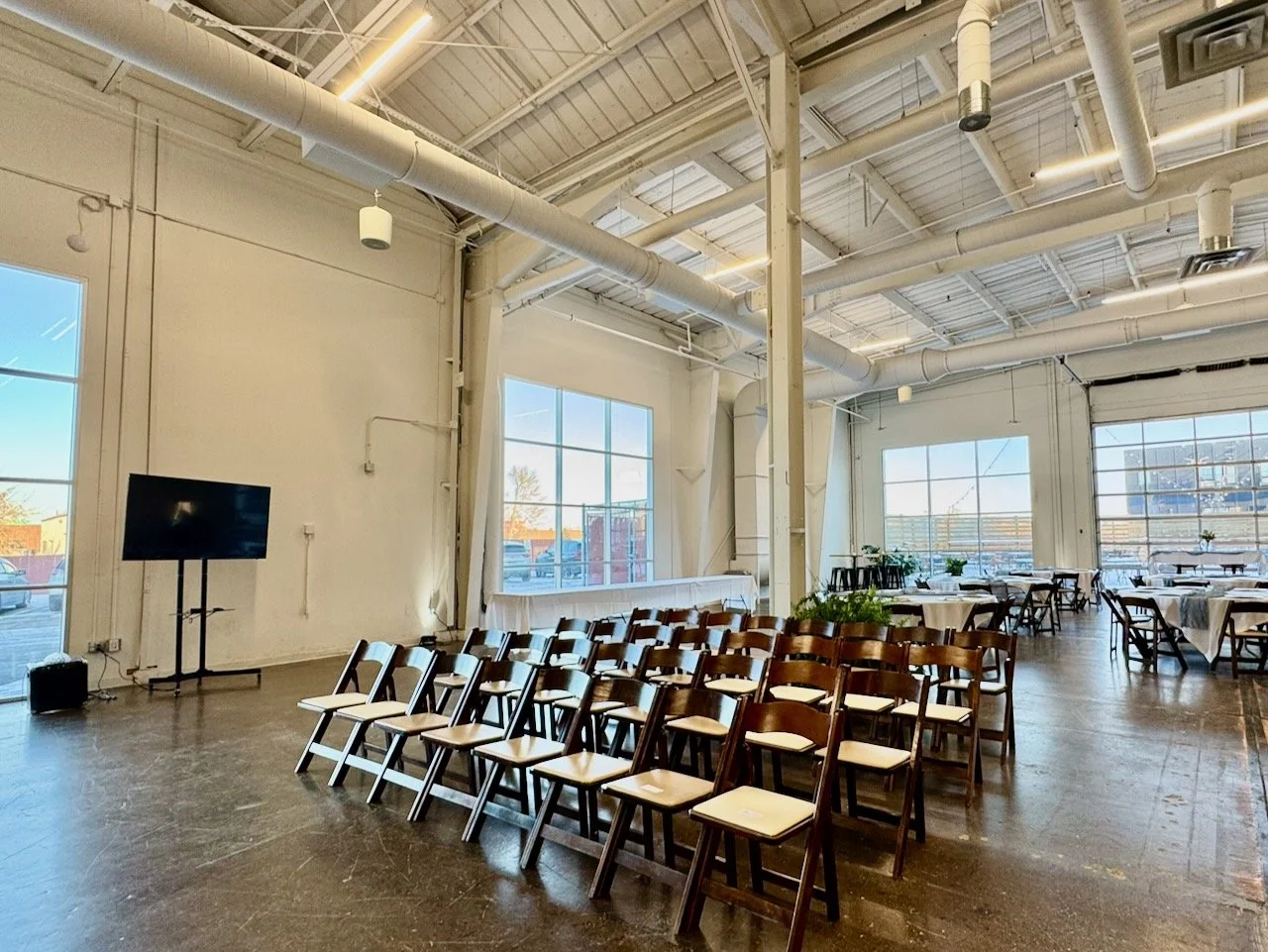 Event space with rows of chairs and round tables, large windows, and a screen on a stand.
