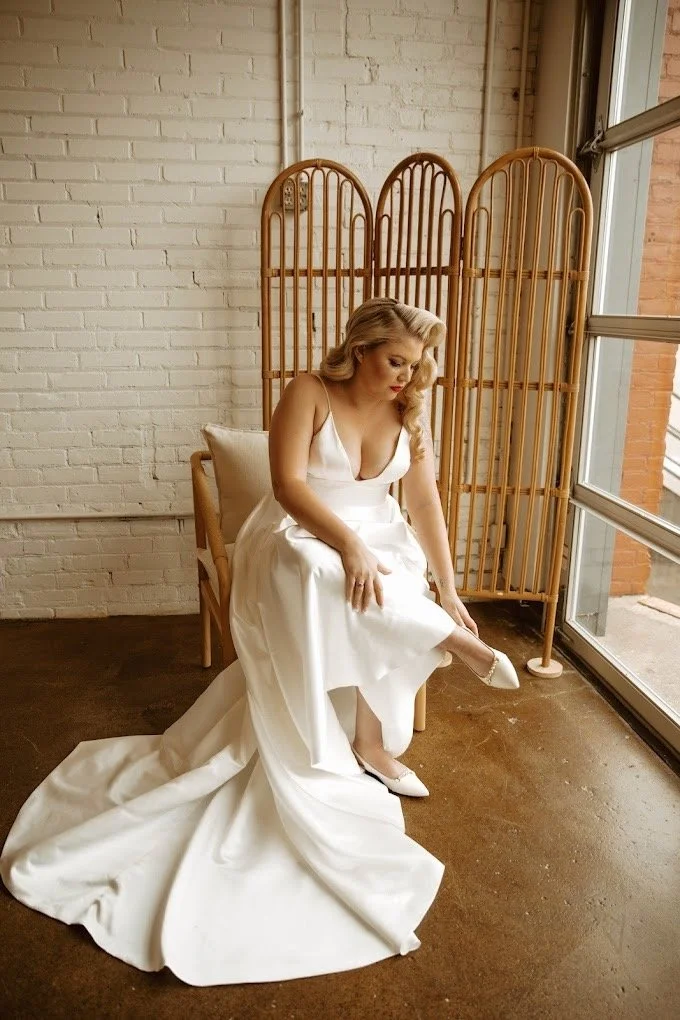 Brunette woman in a white wedding dress sitting on a chair, adjusting her shoe near a large window with daylight and a rattan partition in a room with white brick walls.