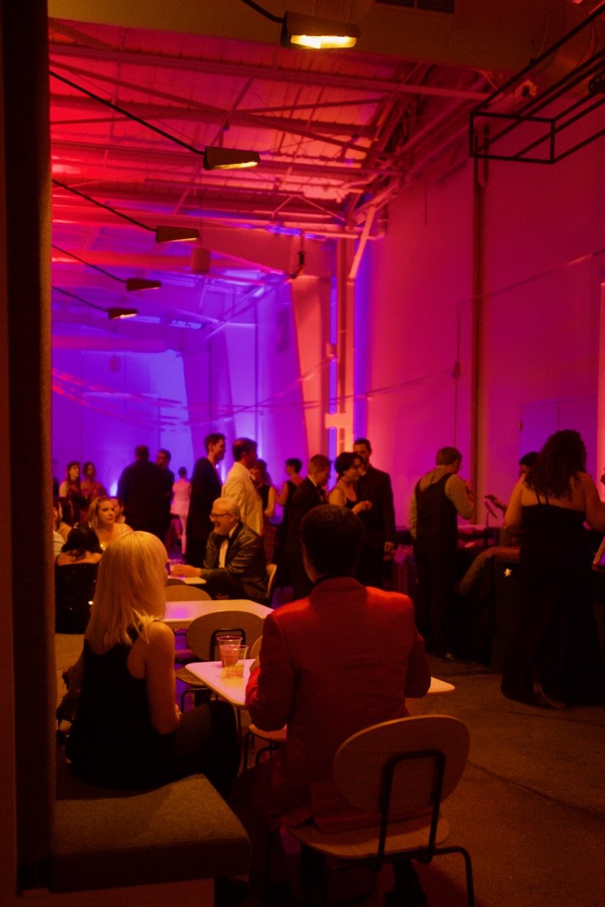 A lively indoor party or social gathering with colorful lighting in shades of pink, purple, and red, featuring people sitting and standing, engaging in conversation, with some seated at tables with drinks and others standing near a bar area.