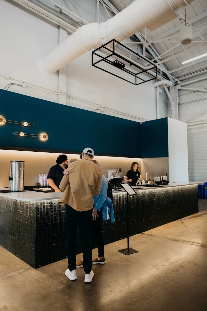 People ordering drinks at a modern coffee shop or bar with a black counter, teal wall, and industrial ceiling.