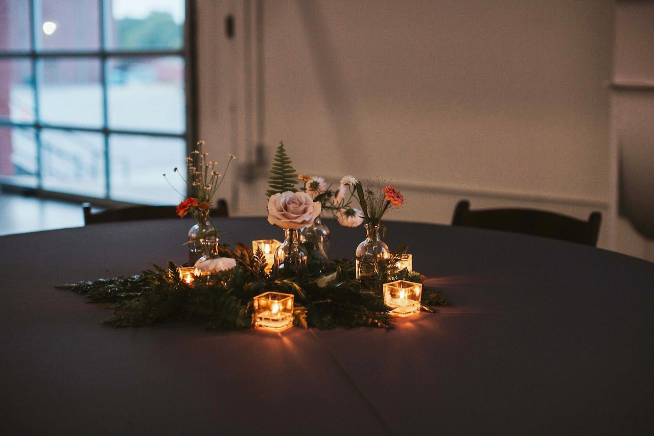 Table centerpiece with flowers in glass vases, surrounded by candles and greenery, in a dimly lit room.