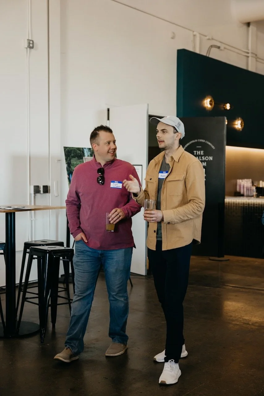 Two men standing and conversing indoors at a social event, each holding a drink, with a bar area and stools nearby.