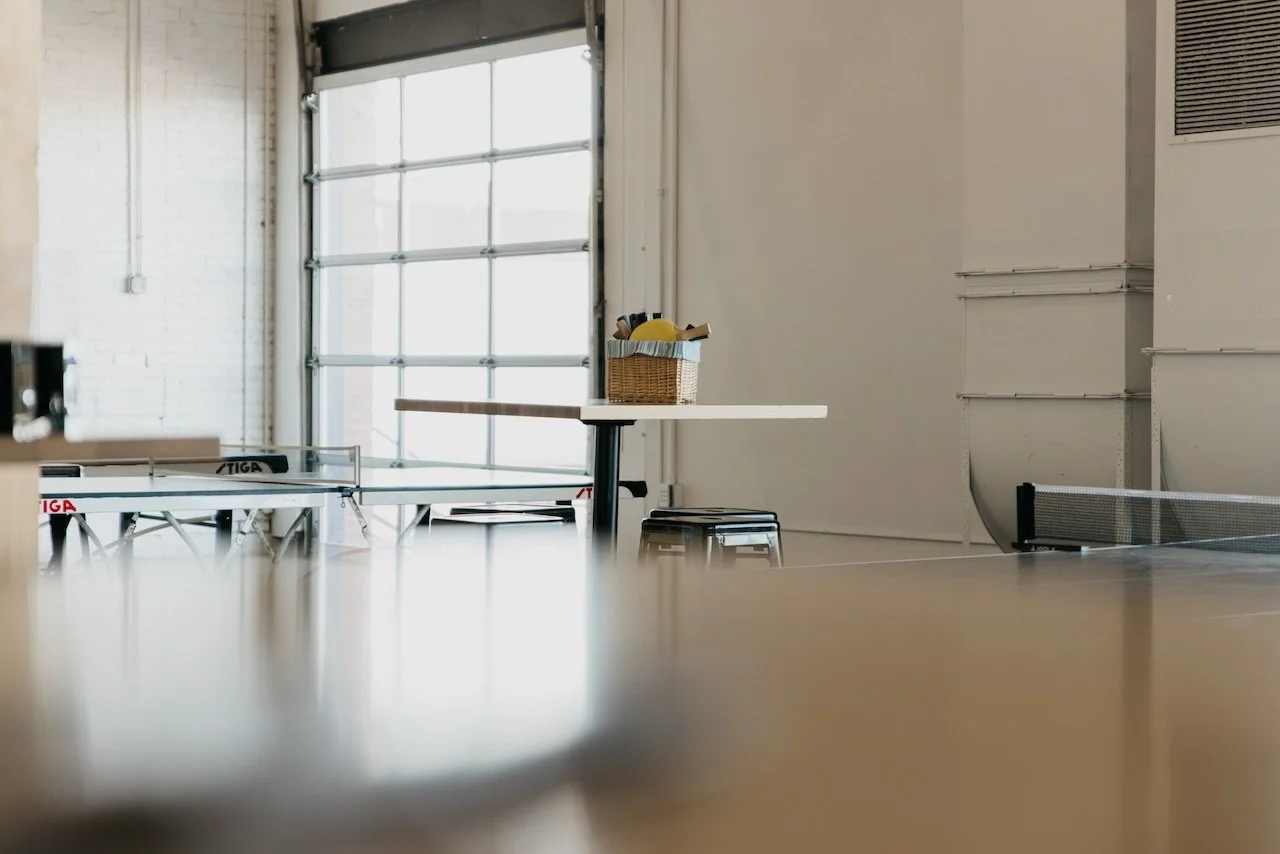 Empty room with ping pong tables, a basket with sports equipment, and a large window letting in natural light.