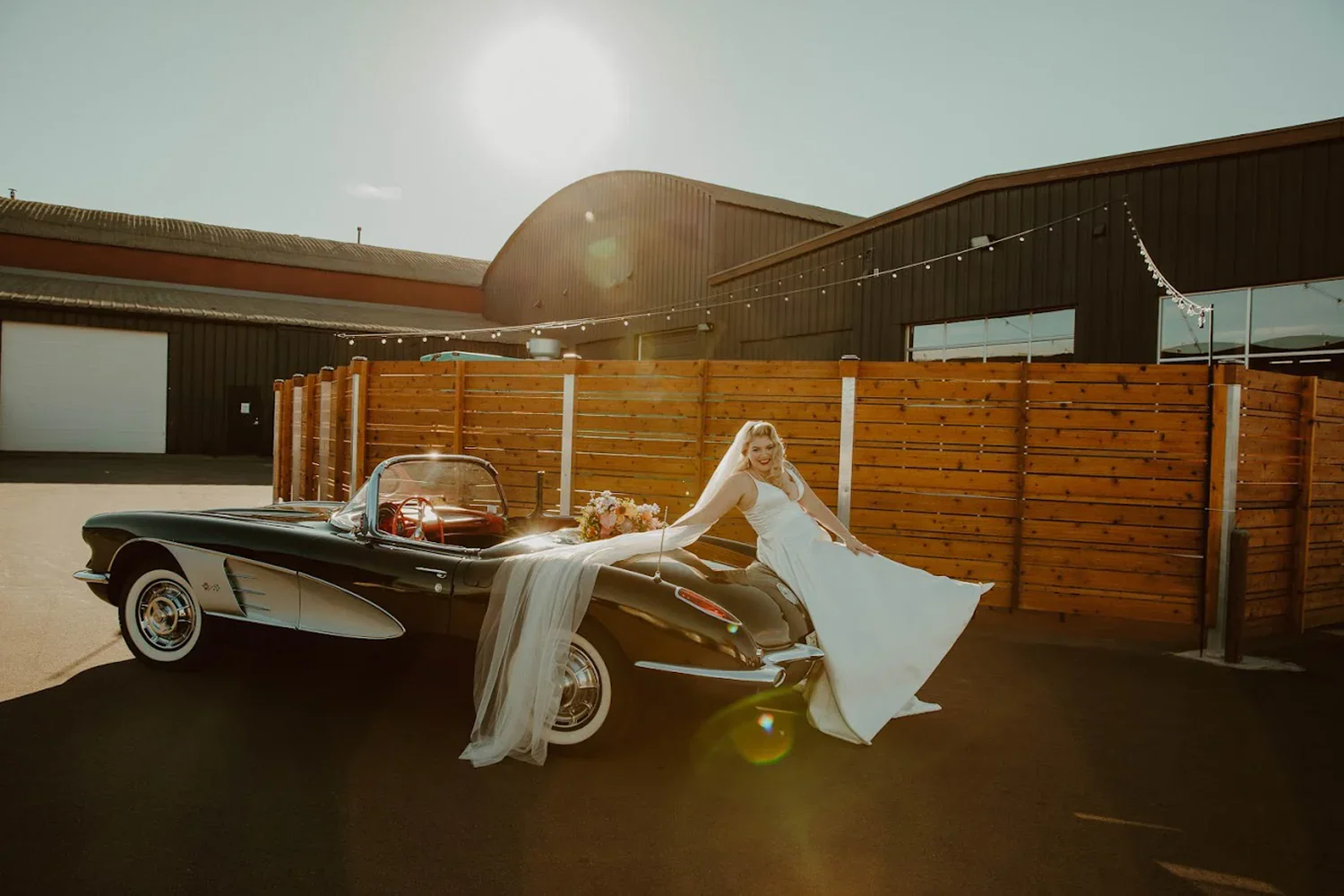 A bride in a white wedding dress is leaning on the hood of a vintage black convertible car, with a bouquet of flowers on the car's hood, in an outdoor setting with a barn and string lights in the background during the daytime.