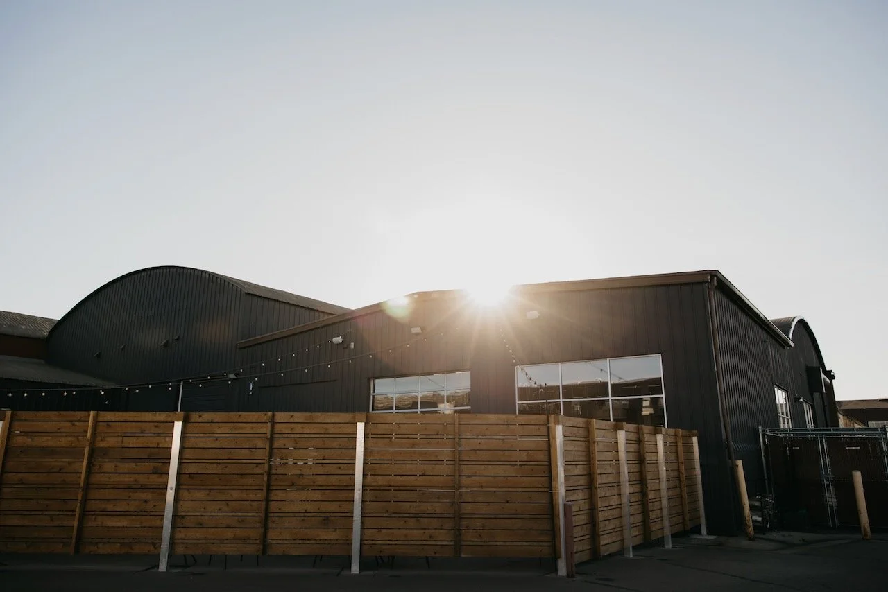 Industrial warehouse with black metal siding and wooden fencing, sunlight shining behind the building.