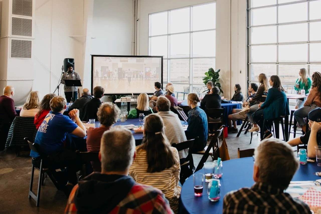 A group of people attending a presentation in a bright, spacious room with large windows. The presenter stands at a podium with a large screen displaying a video or slides behind him. Attendees are seated at round tables, listening attentively.