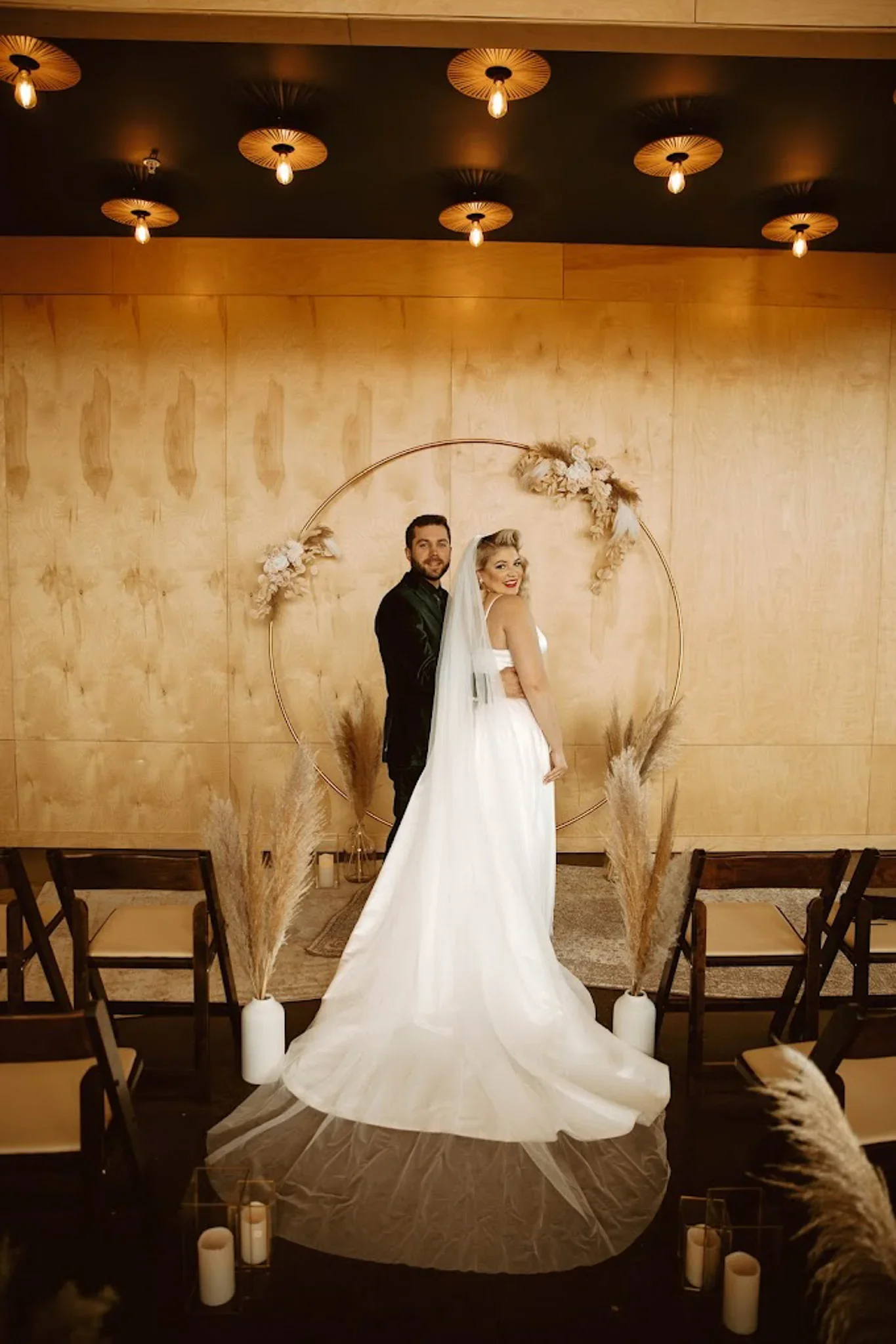A bride and groom standing together, smiling, in front of a decorative circle with dried floral arrangements, on a wooden backdrop, with candles and pampas grass in vases around them, at a wedding ceremony.