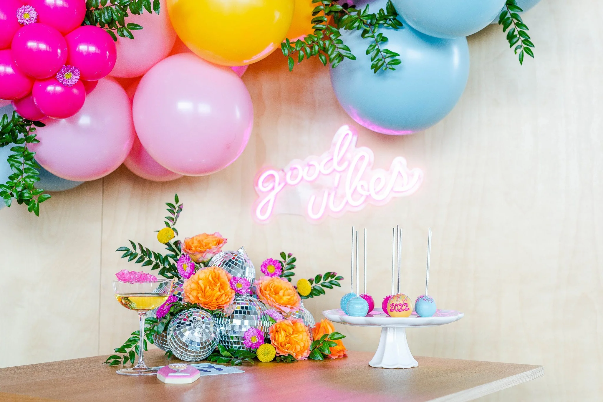 Decorated table with disco balls, orange and pink flowers, a cocktail glass, and pink and blue cake pops, with pink and blue balloons and a neon sign that says 'good vibes' in the background.