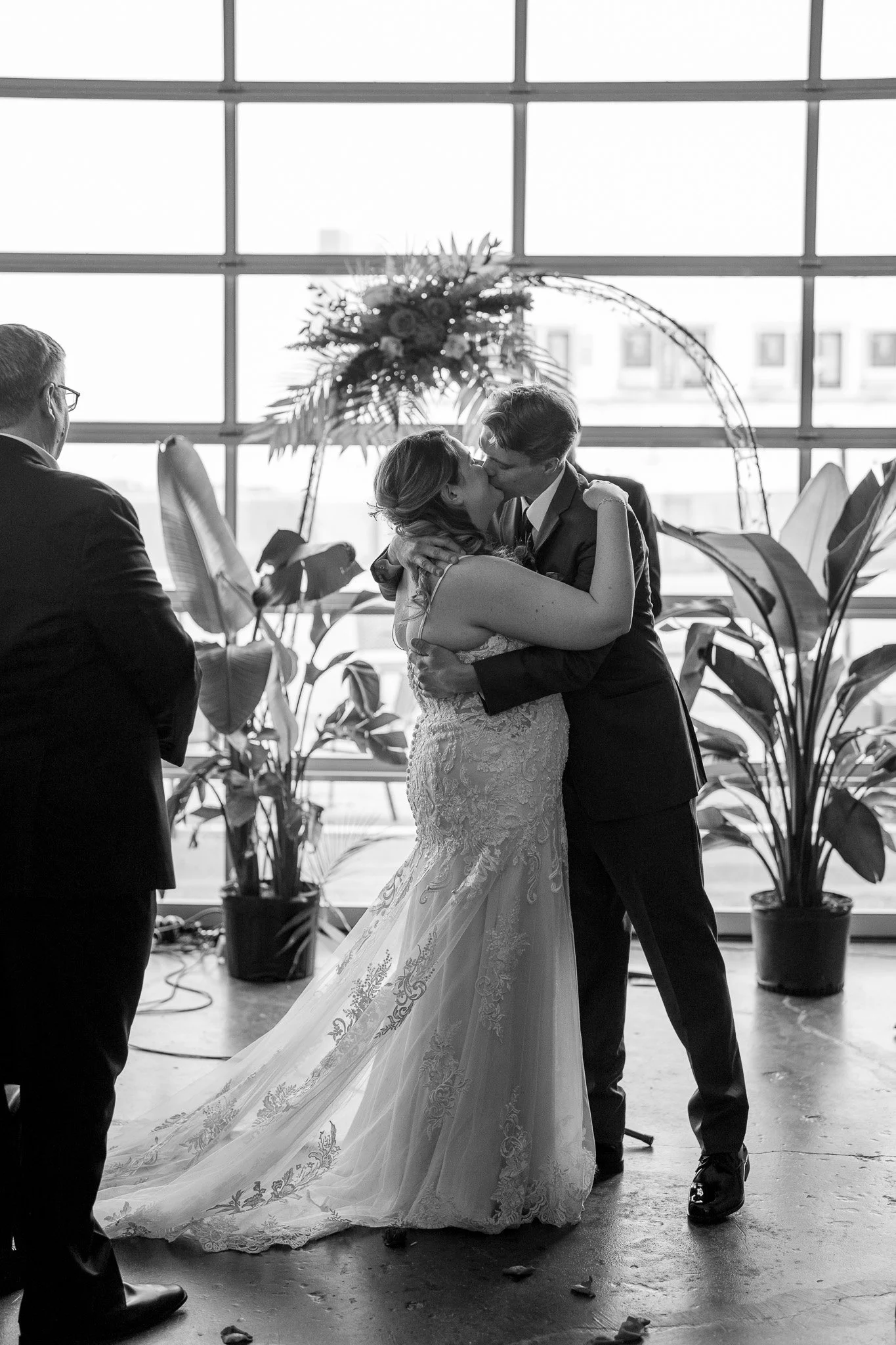 A couple sharing a kiss at their wedding ceremony with a man standing nearby, inside a venue with large windows and plants in the background.