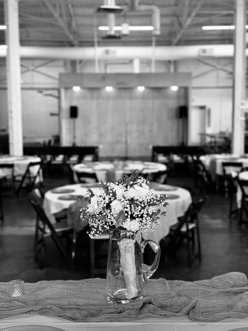 A floral centerpiece in a glass pitcher on a table in a decorated event venue with round tables and chairs, and a stage with speakers in the background.