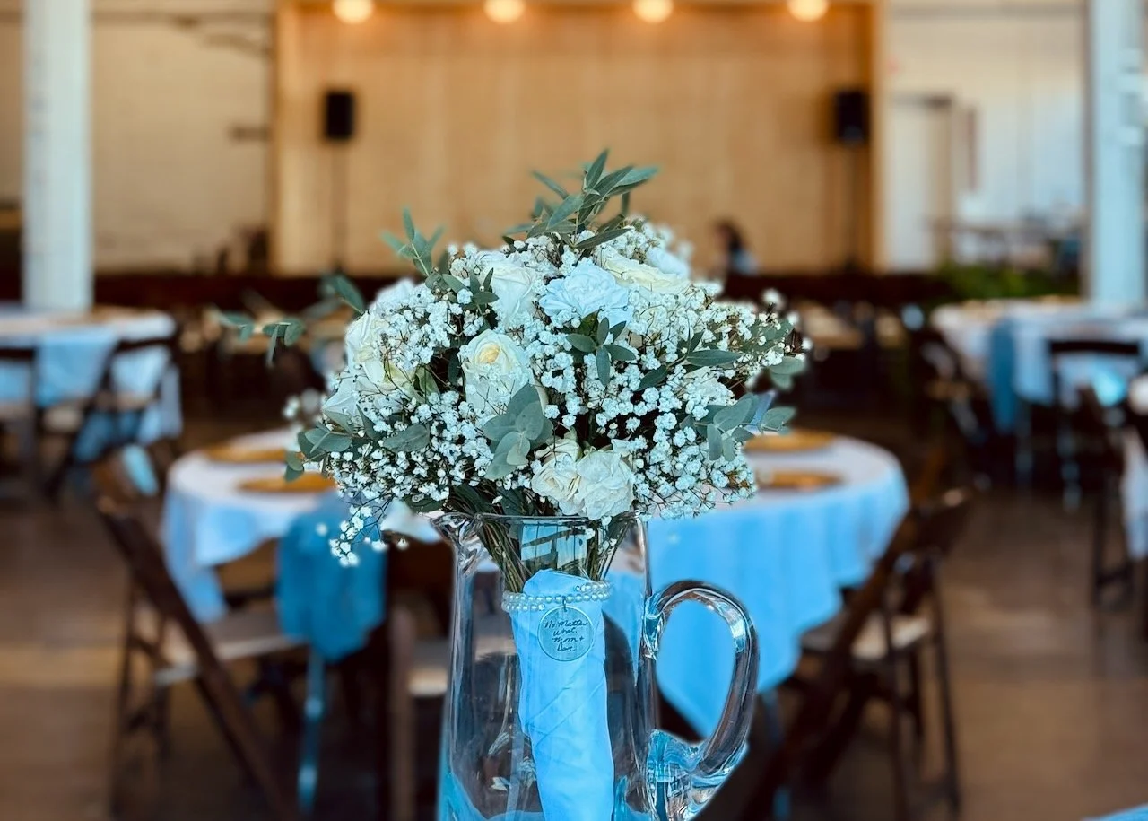 A floral centerpiece with white roses, baby's breath, and eucalyptus in a clear glass pitcher on a table in a decorated event hall.