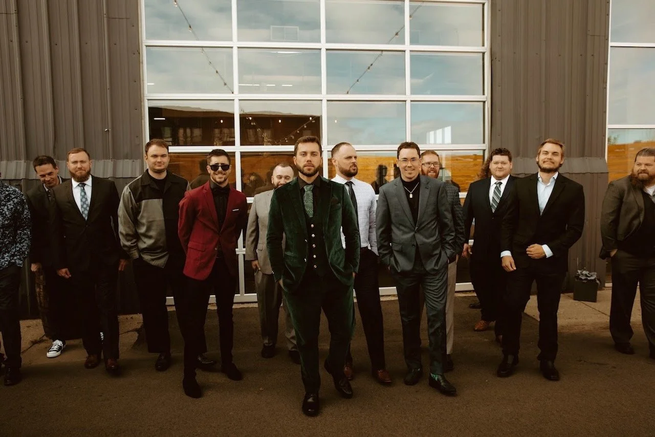 Group of men dressed in formal and semi-formal attire standing outside a building with large glass windows.