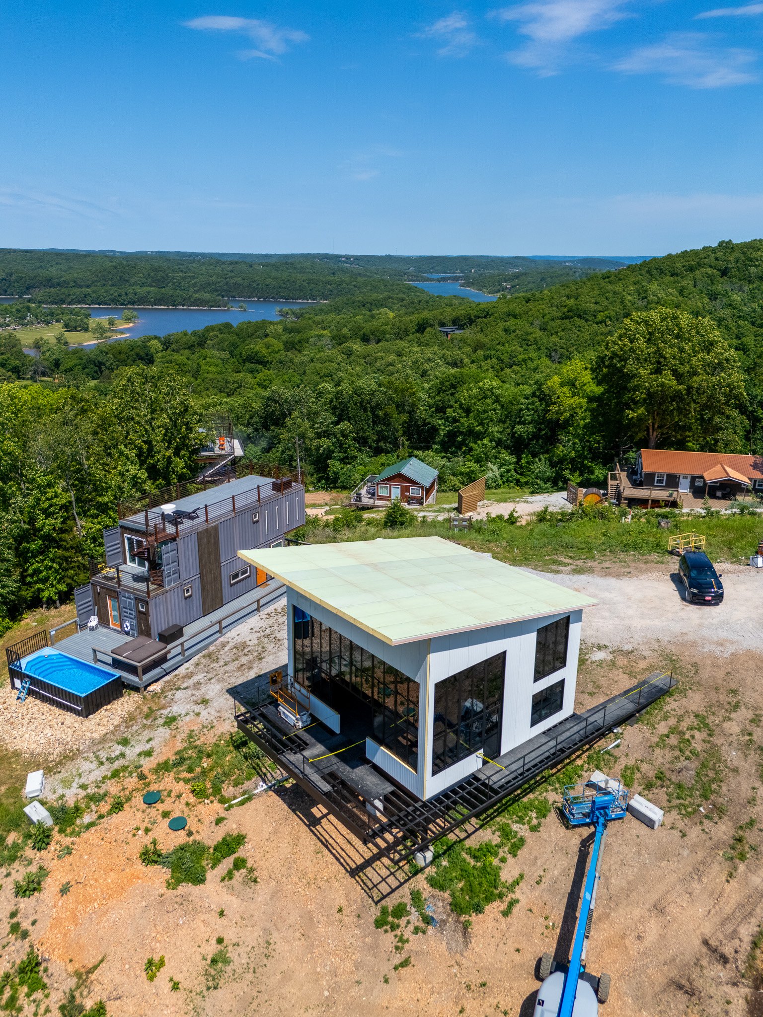 Aerial view of a construction site with a modern white house, construction equipment, and surrounding greenery near a body of water and hills.