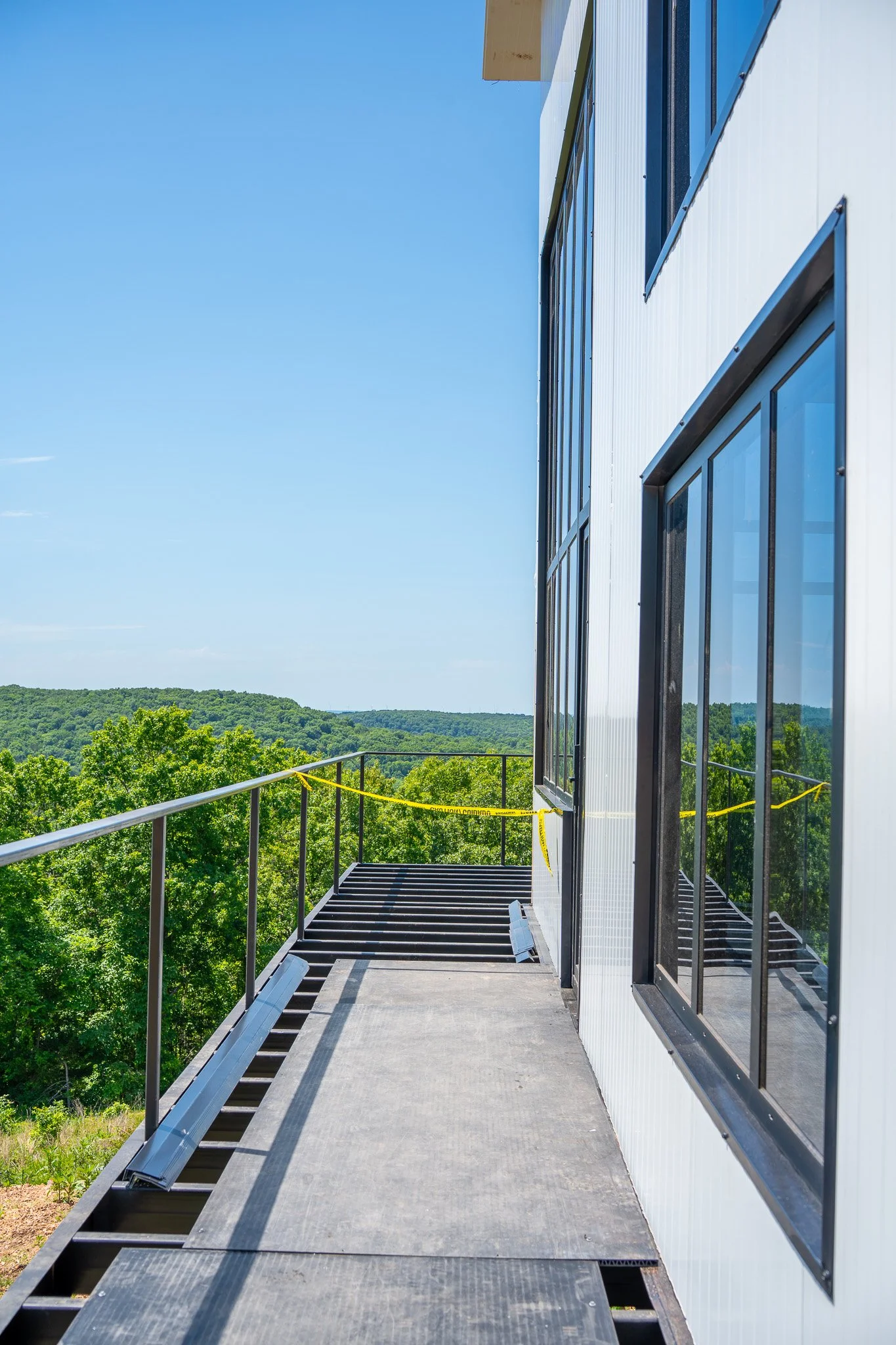 Outdoor balcony or fire escape with metal railing and grated floor, attached to a modern building with large windows and white exterior walls, overlooking a lush green landscape with trees and hills under a clear blue sky.