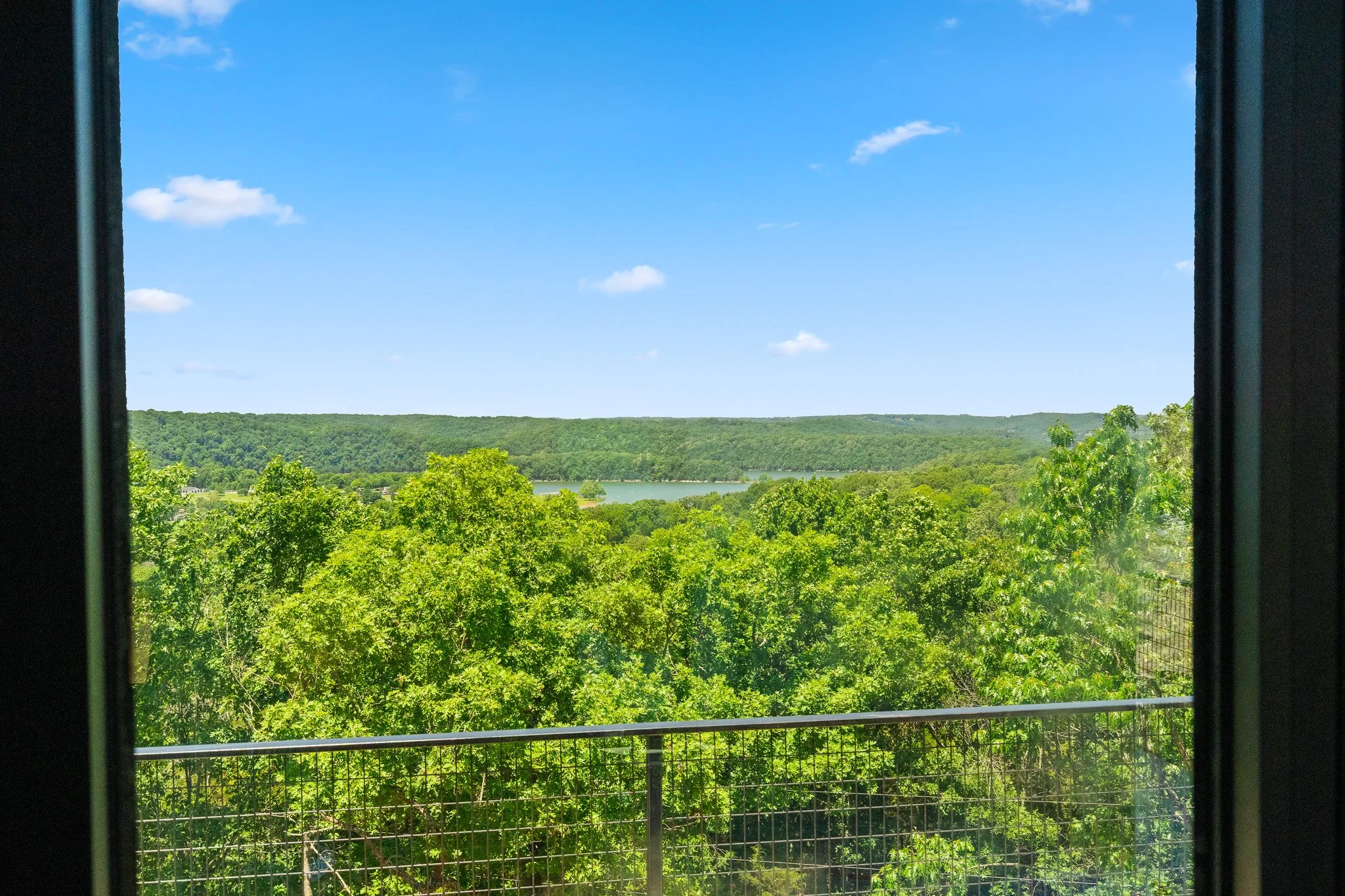 View of a green landscape with trees, a lake, and hills under a blue sky with scattered clouds, seen through a window with black framing and a metal railing in the foreground.