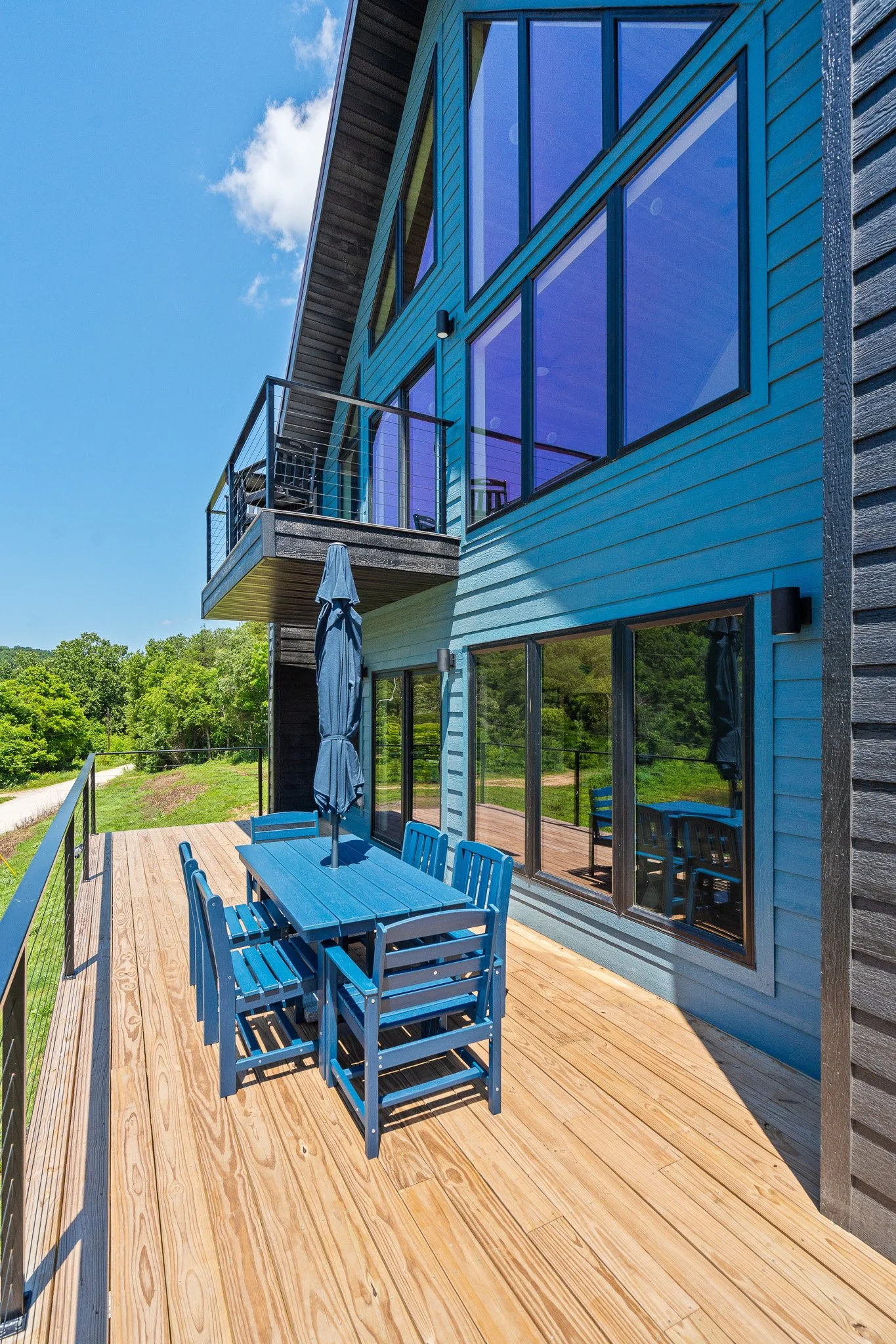 Modern blue house with a wooden deck and glass windows and doors, outdoor seating area with blue table and chairs, a closed umbrella, and trees in the background under a partly cloudy sky.