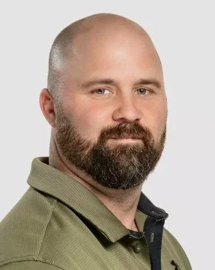 A portrait of a bald man with a beard, wearing a green collared shirt, against a plain light background.