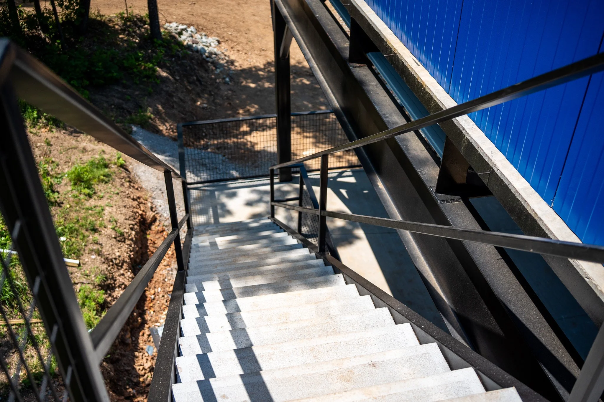 Exterior view of a staircase with black railings and white steps, leading down to a dirt and grass area. A blue building wall with vertical siding is visible on the right. The staircase is partially shaded by nearby trees.
