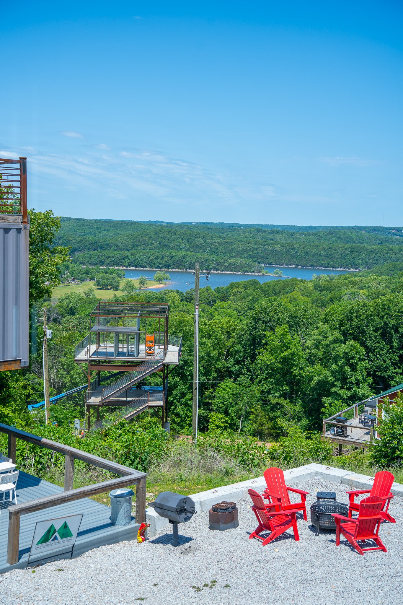 A backyard patio with a firepit surrounded by four red Adirondack chairs, outdoor grill, and gravel ground, overlooking a scenic view of lush green trees, a river, and rolling hills under a clear blue sky.